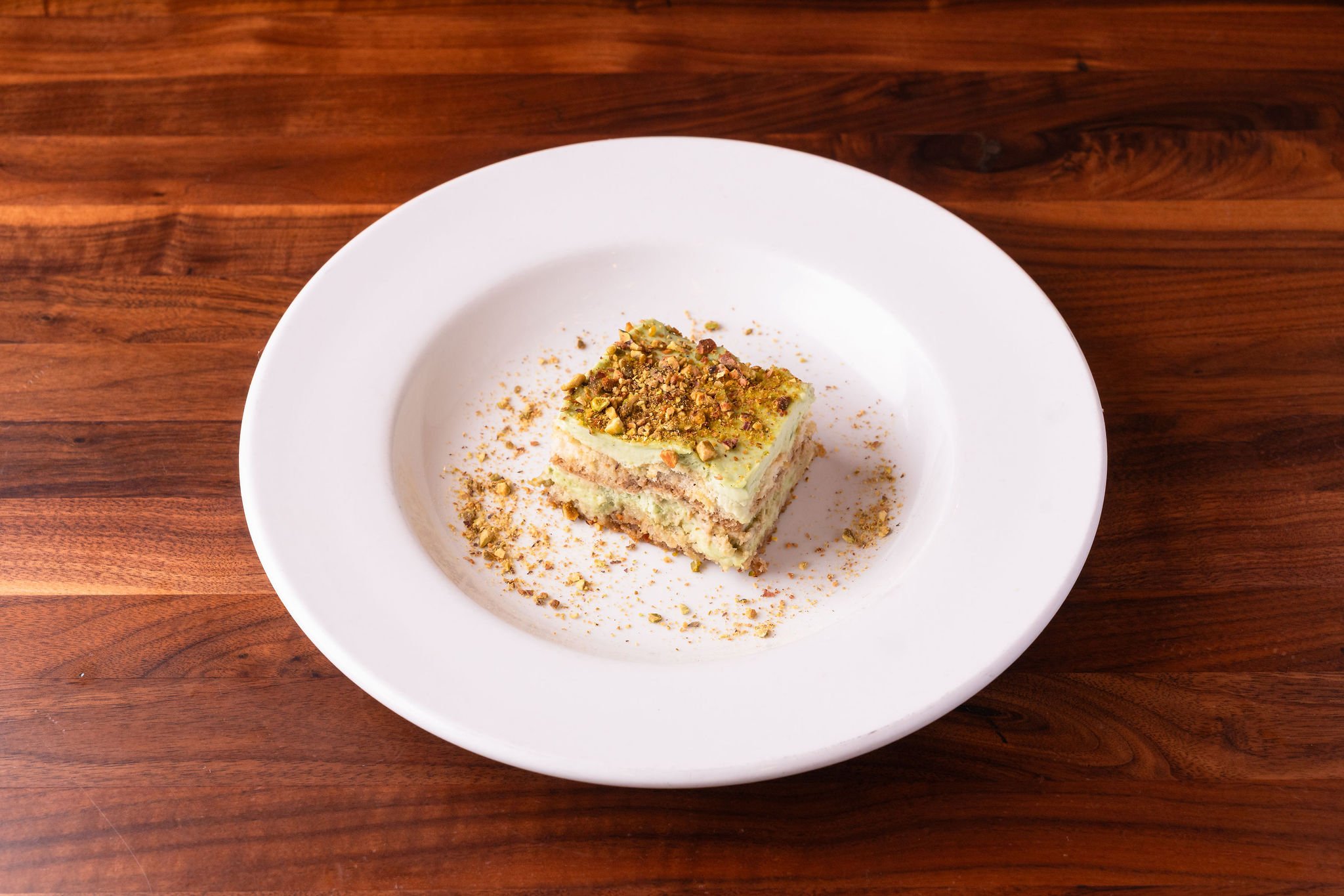A slice of layered pistachio dessert with crushed pistachios on top on a white plate placed on a wooden table.