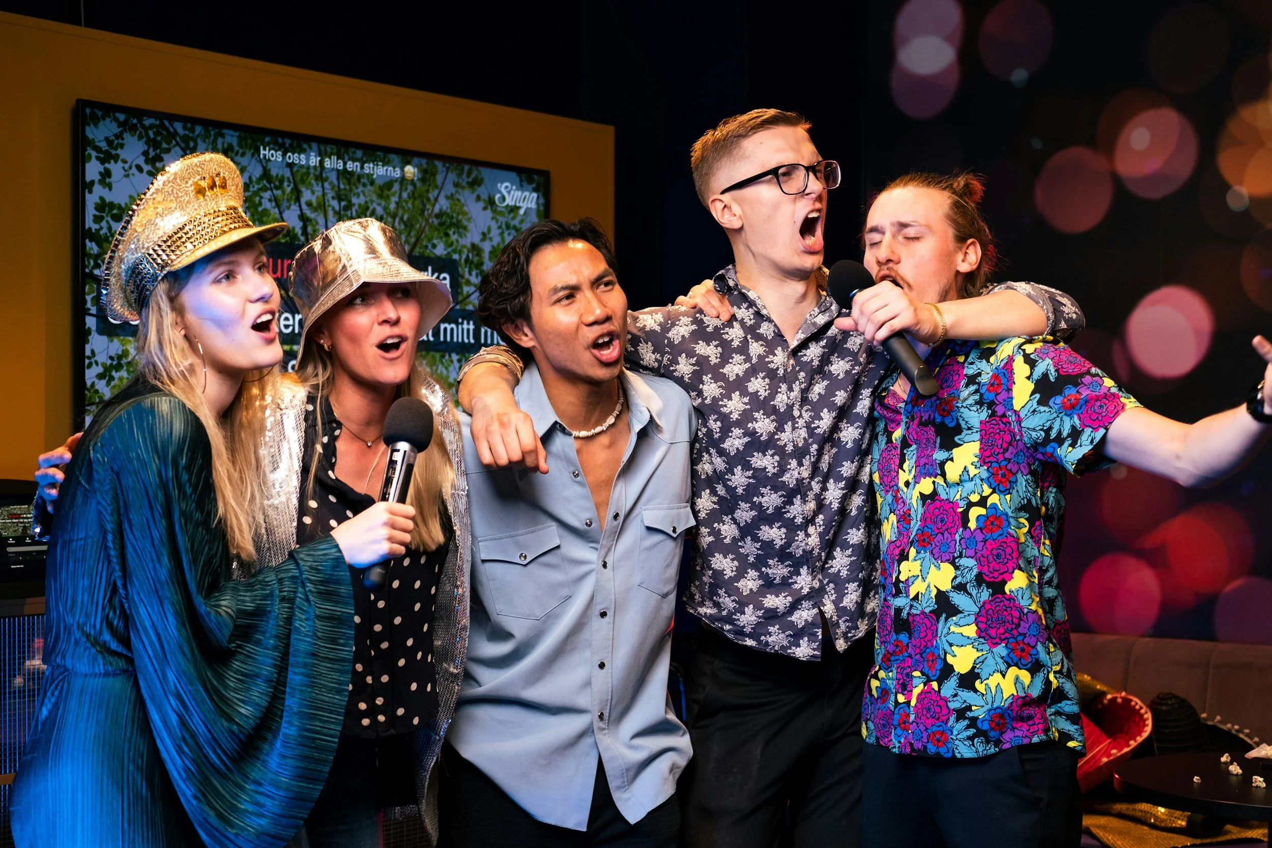 Five young adults singing together on stage, holding microphones, with a background of colorful lights and a TV screen.