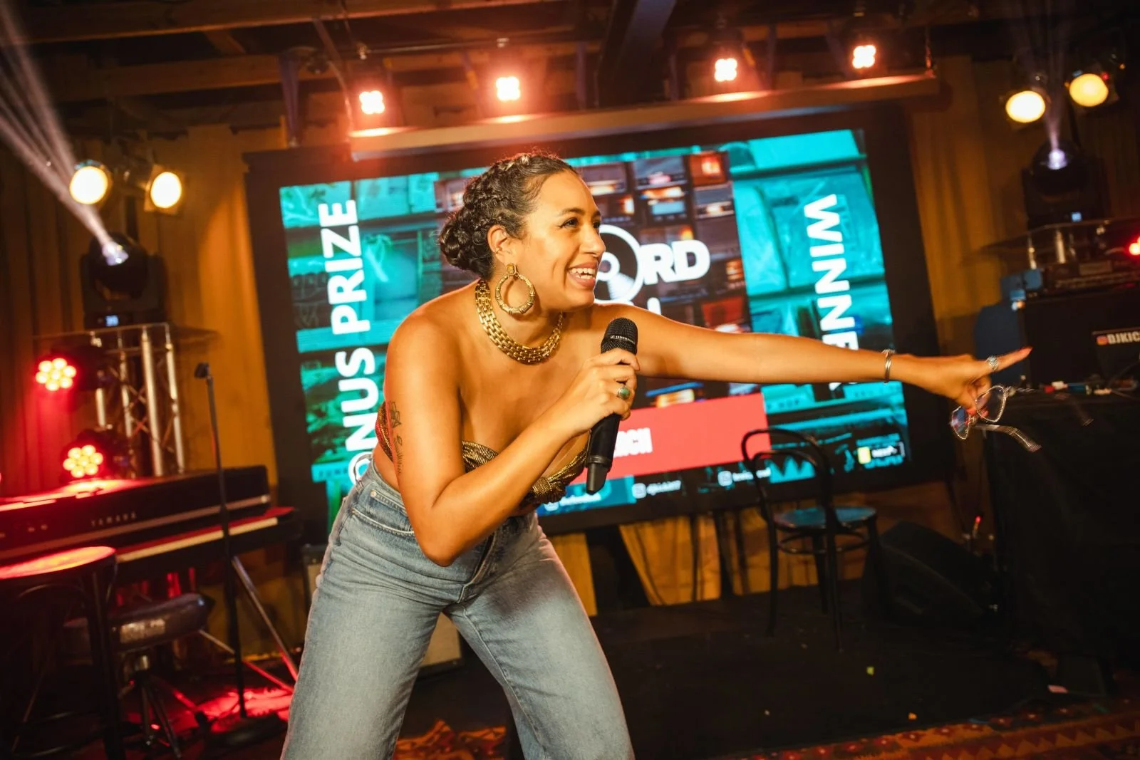 A woman with curly hair styled in an updo, wearing gold jewelry, gesturing and speaking into a microphone on stage at an event with a large screen displaying words in the background.