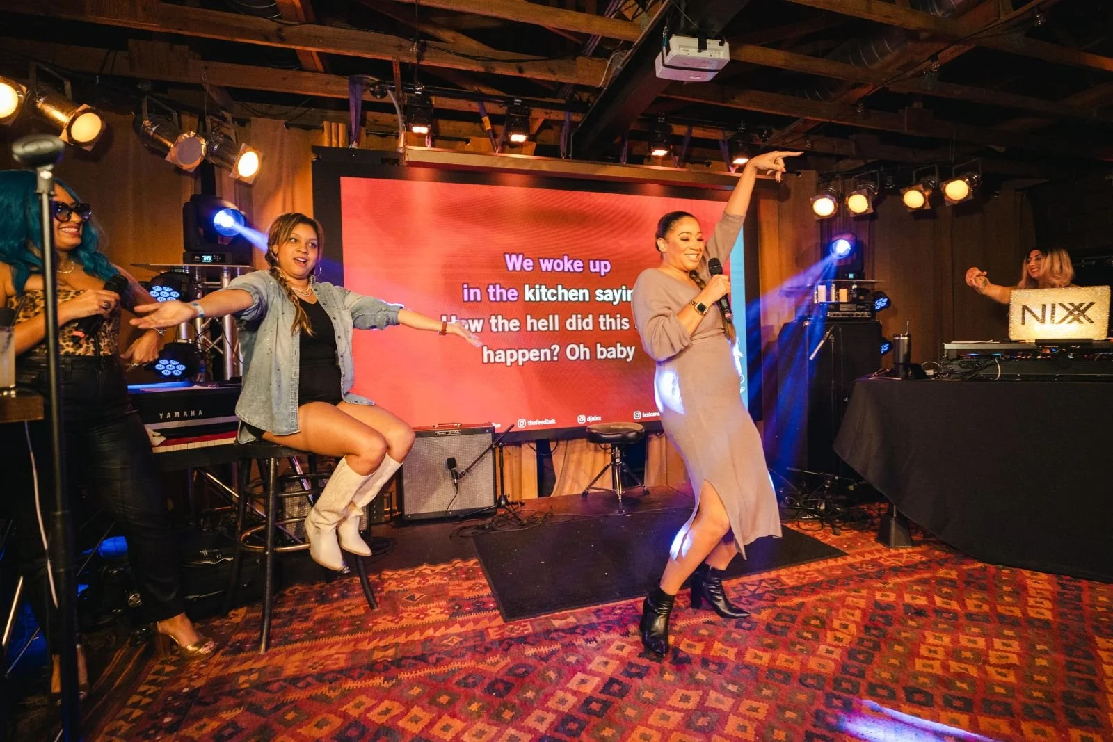 Women performing on stage at karaoke night with large screen displaying song lyrics behind them.