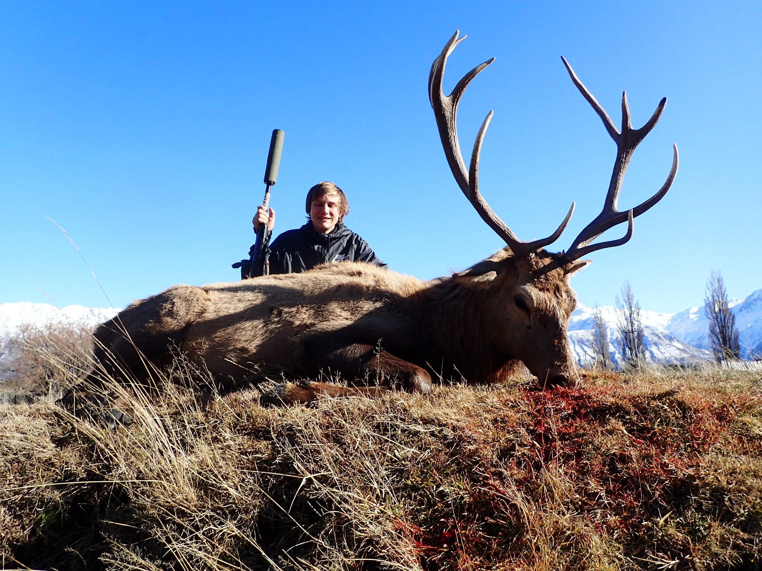 A young man with blonde hair holding a rifle, sitting behind a large, dead elk with impressive antlers, on a grassy hillside with mountains and snow in the background.