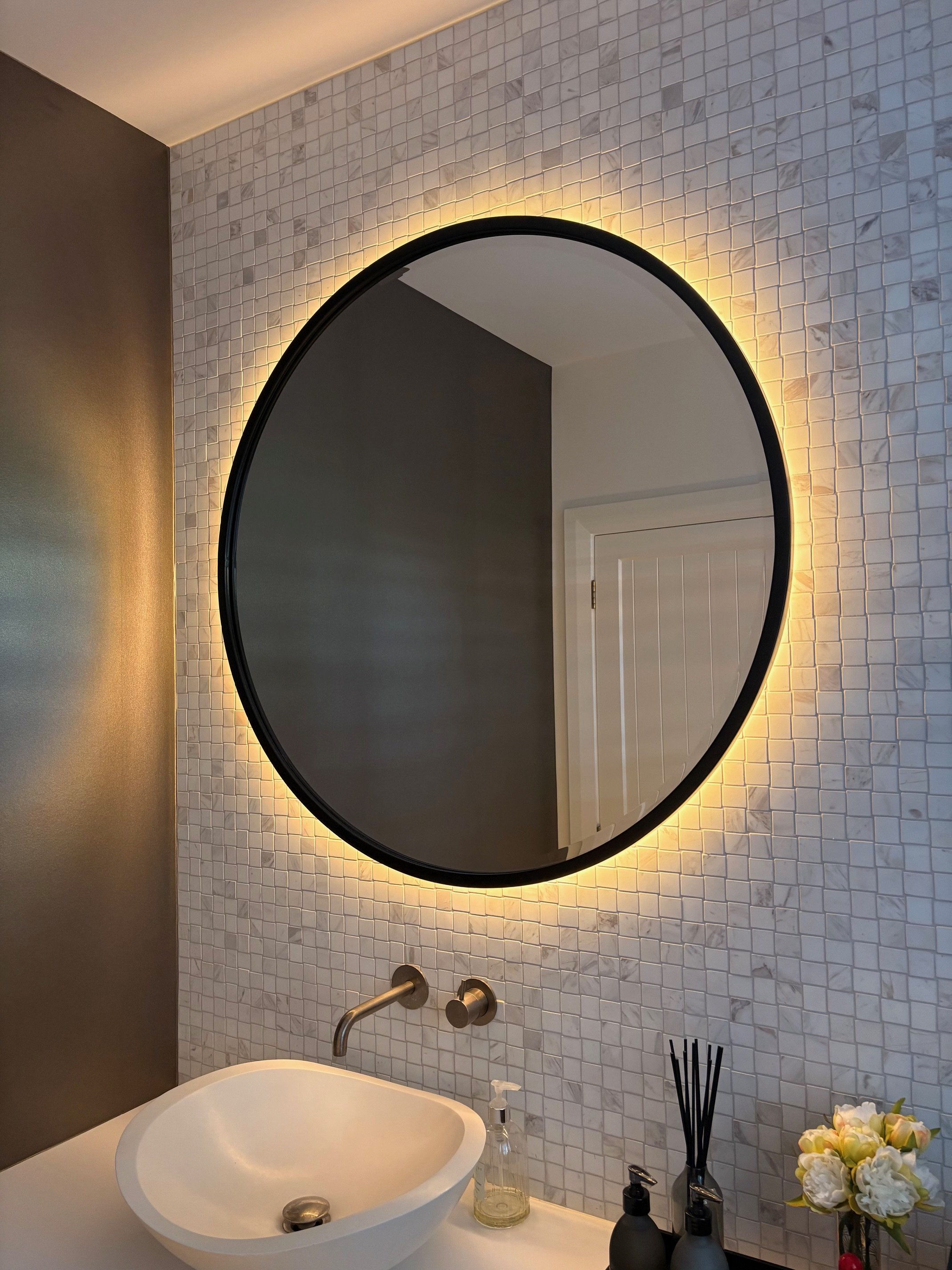 Modern bathroom vanity with a white vessel sink, a wall-mounted faucet, soap dispenser, reed diffuser, black and white bottles, and a mirror with backlighting, set against a textured tiled wall.