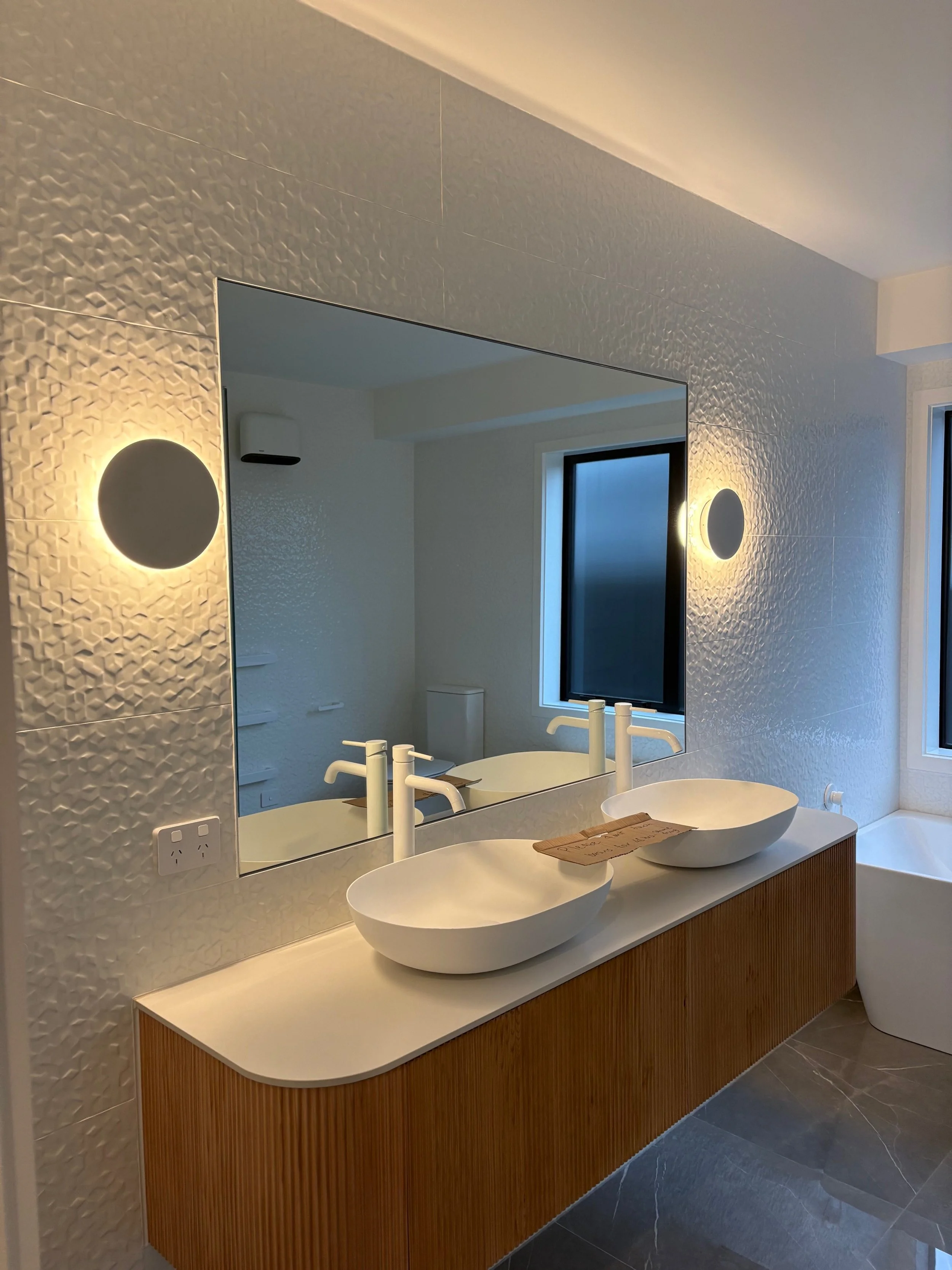 Modern bathroom with textured white walls, large mirror, two bowl sinks with white faucets on a wooden vanity, circular wall lights, a window, and a bathtub.