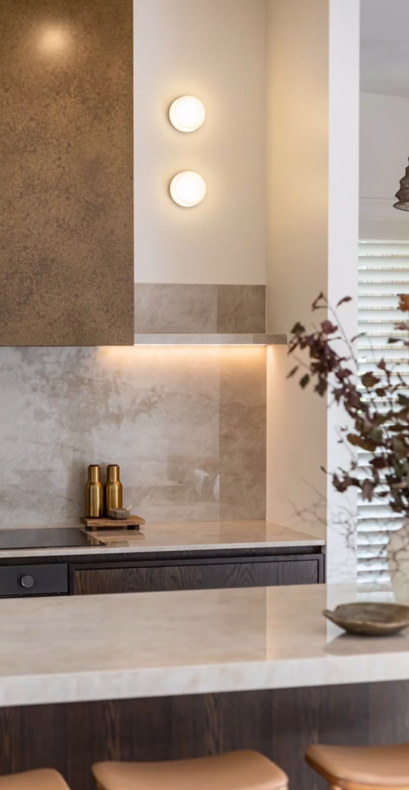 Modern kitchen with beige countertops, dark wood cabinets, and decorative gold bottles on a tray. Wall has two round wall sconces and a textured accent wall. Part of a window with blinds and a vase with dried flowers are visible.