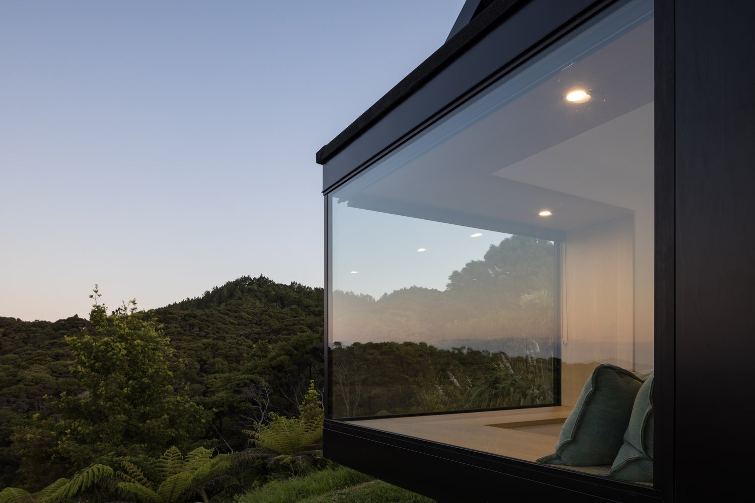 Modern glass house with a black frame overlooking a lush green hillside during dusk.