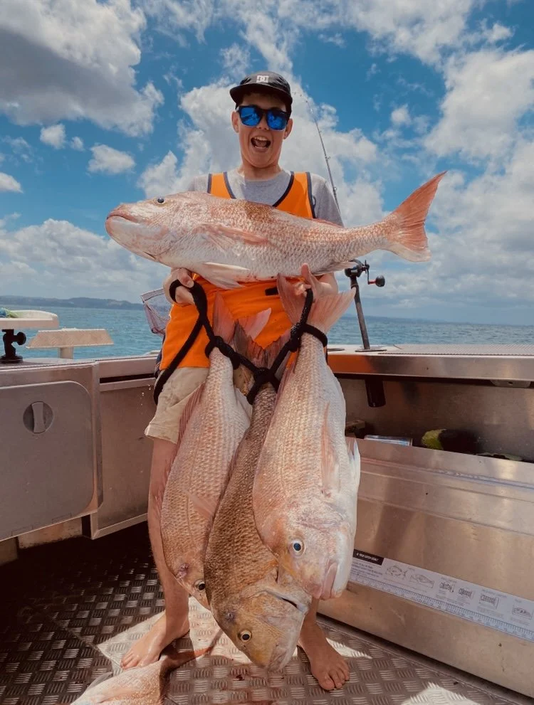 A person on a boat holding a large fish with three other fish hanging down from their arms, with a blue sky and ocean in the background.