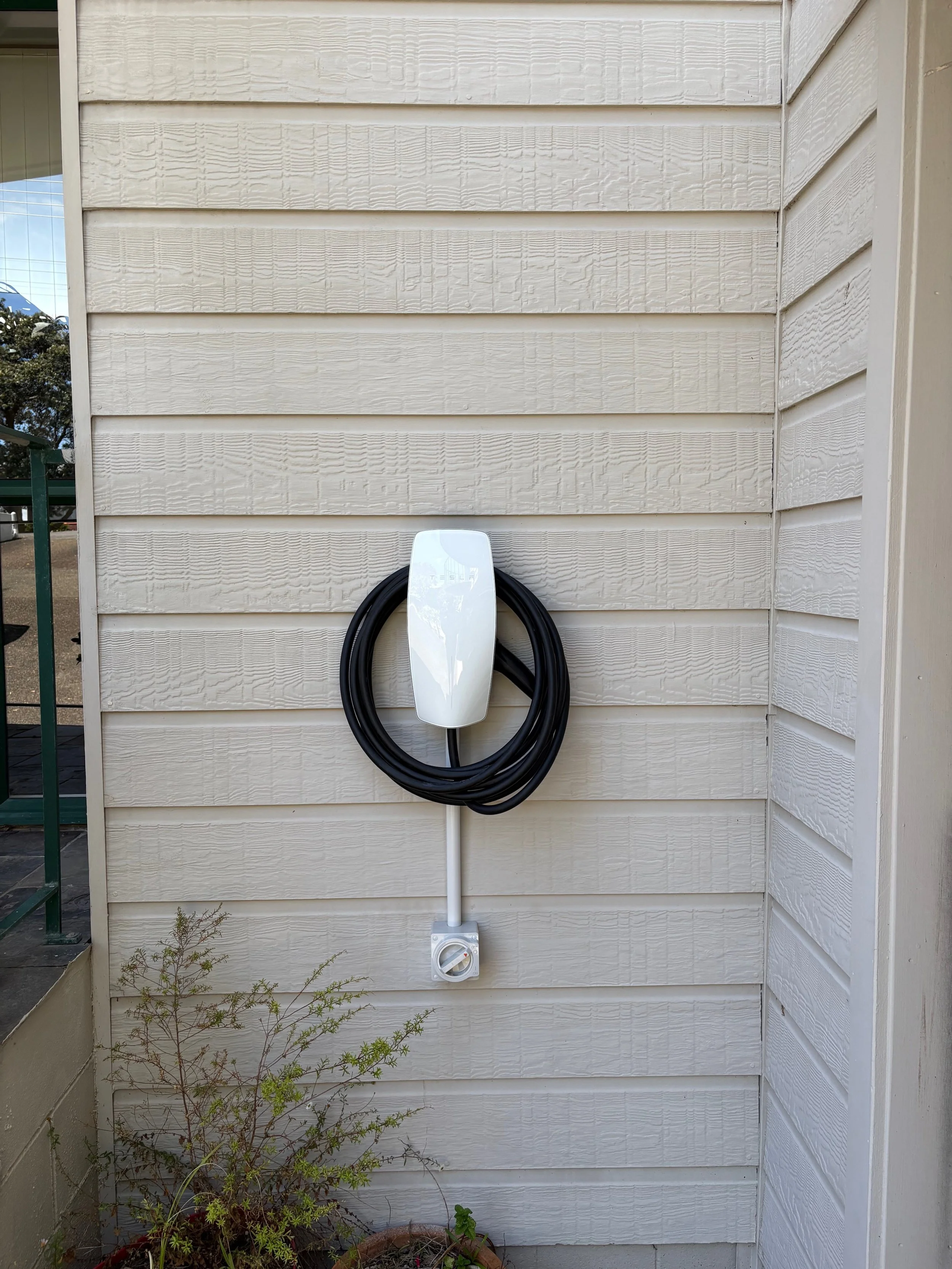 Electric vehicle charging station with a white cover, black cable coiled around it, and a power outlet at the bottom, mounted on a beige wooden wall outside.