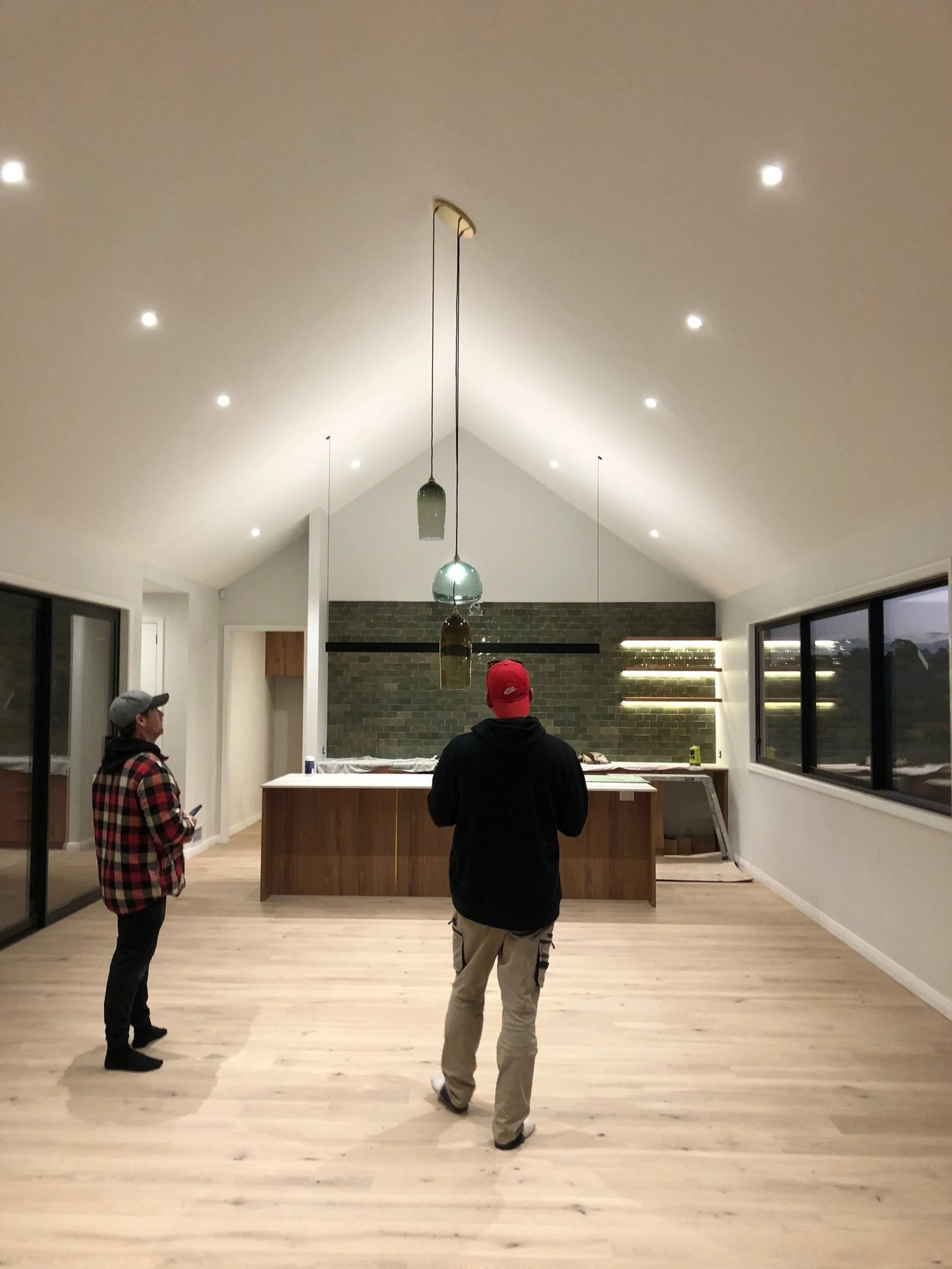Two men standing in a modern, minimally decorated room with high vaulted ceiling, pendant lighting, large windows, and a kitchen island at the back, appearing to be inspecting the space.