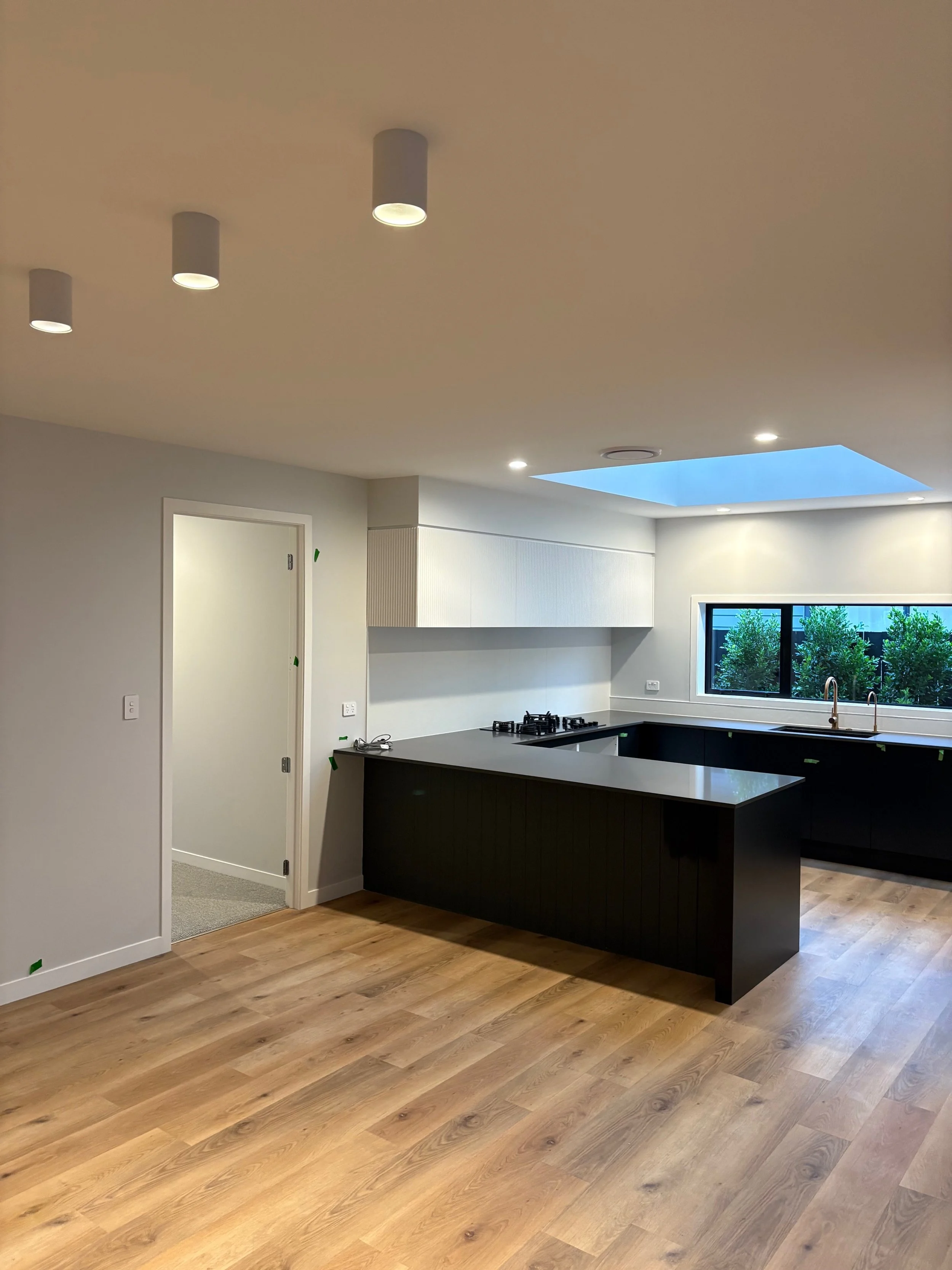 Modern kitchen with black cabinets, wooden floor, white upper cabinets, window with greenery, and a skylight ceiling.