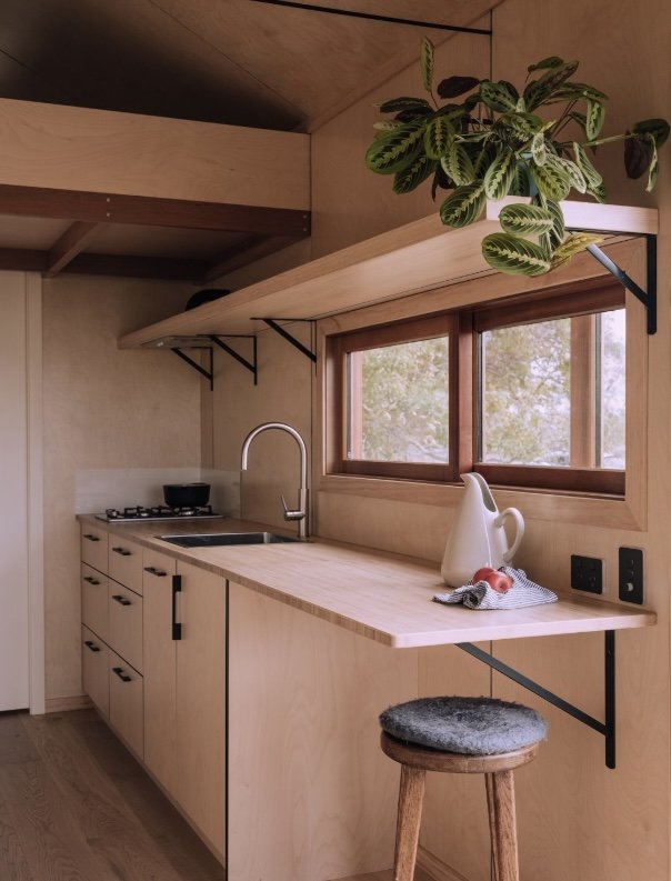 A minimalist kitchen with light wood cabinetry, a small sink, a gas stove, a large window, and a wooden counter with a teapot, a fruit, and a cloth. There is a wooden stool with a cushioned seat and a potted plant on a floating shelf above the window