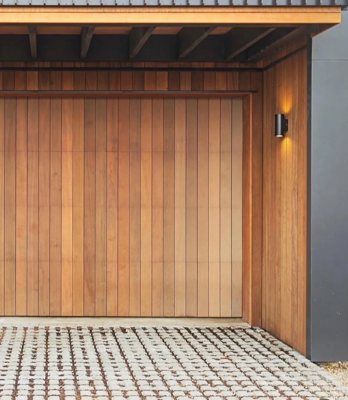 Wooden garage door with a modern wall light on the right side, next to a stone or brick paved driveway.