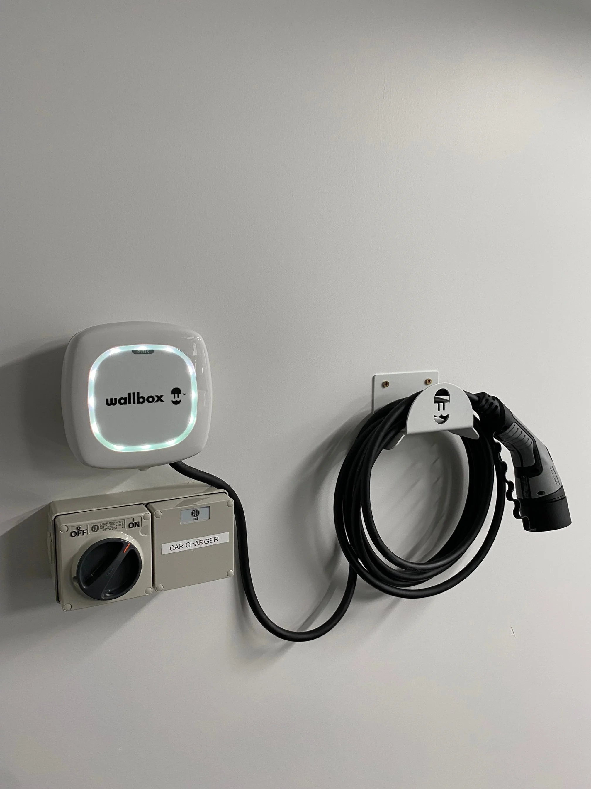 Electric vehicle wall charging station with a Wallbox charger, a power outlet labeled 'Car Charger,' and an electric vehicle charging cable with a plug hanging on a wall.