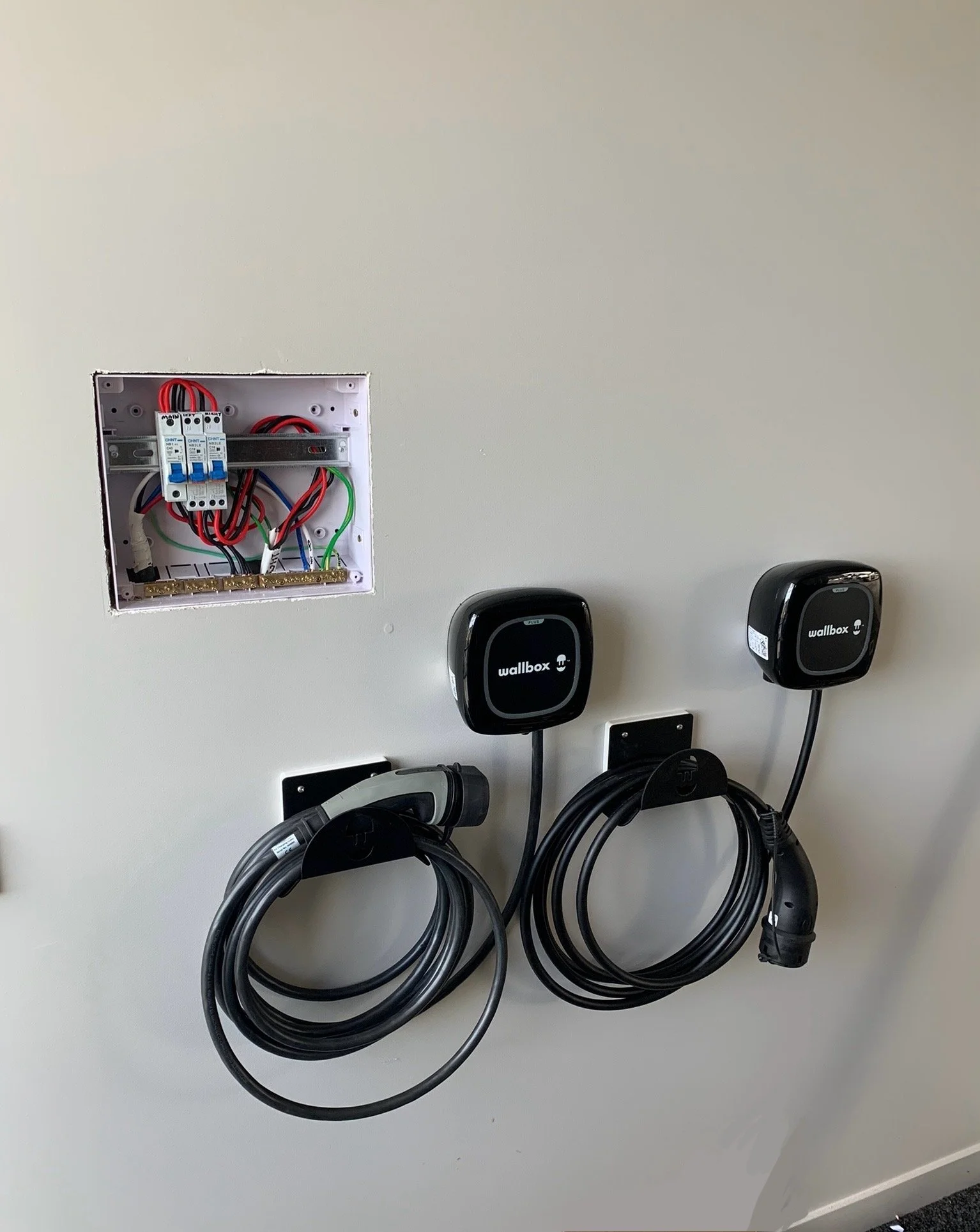 Electrical panel with exposed wiring and two Wallbox electric vehicle charging stations mounted on a white wall.