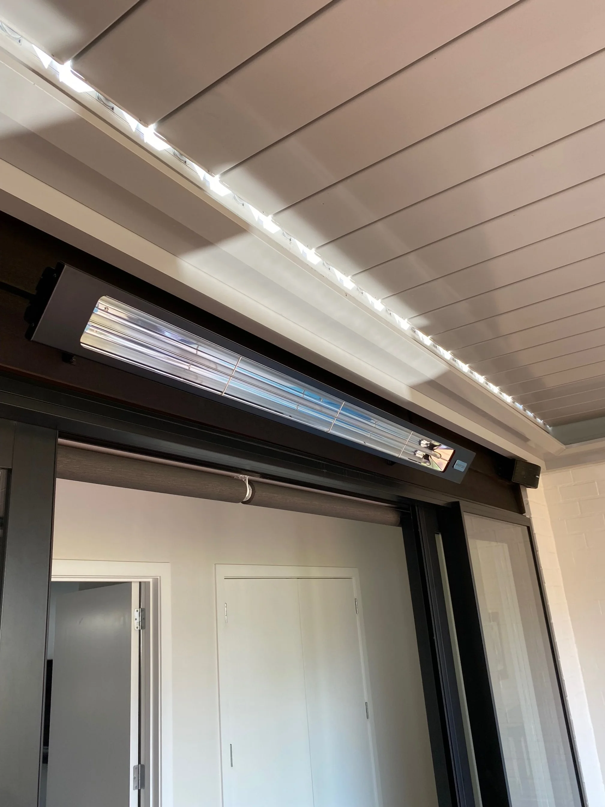 The image shows a close-up view of a modern sliding glass door with black aluminum frames, a gray roller shade partially rolled up, and a white ceiling with an automated window blind system that has a metallic and black casing. The background include