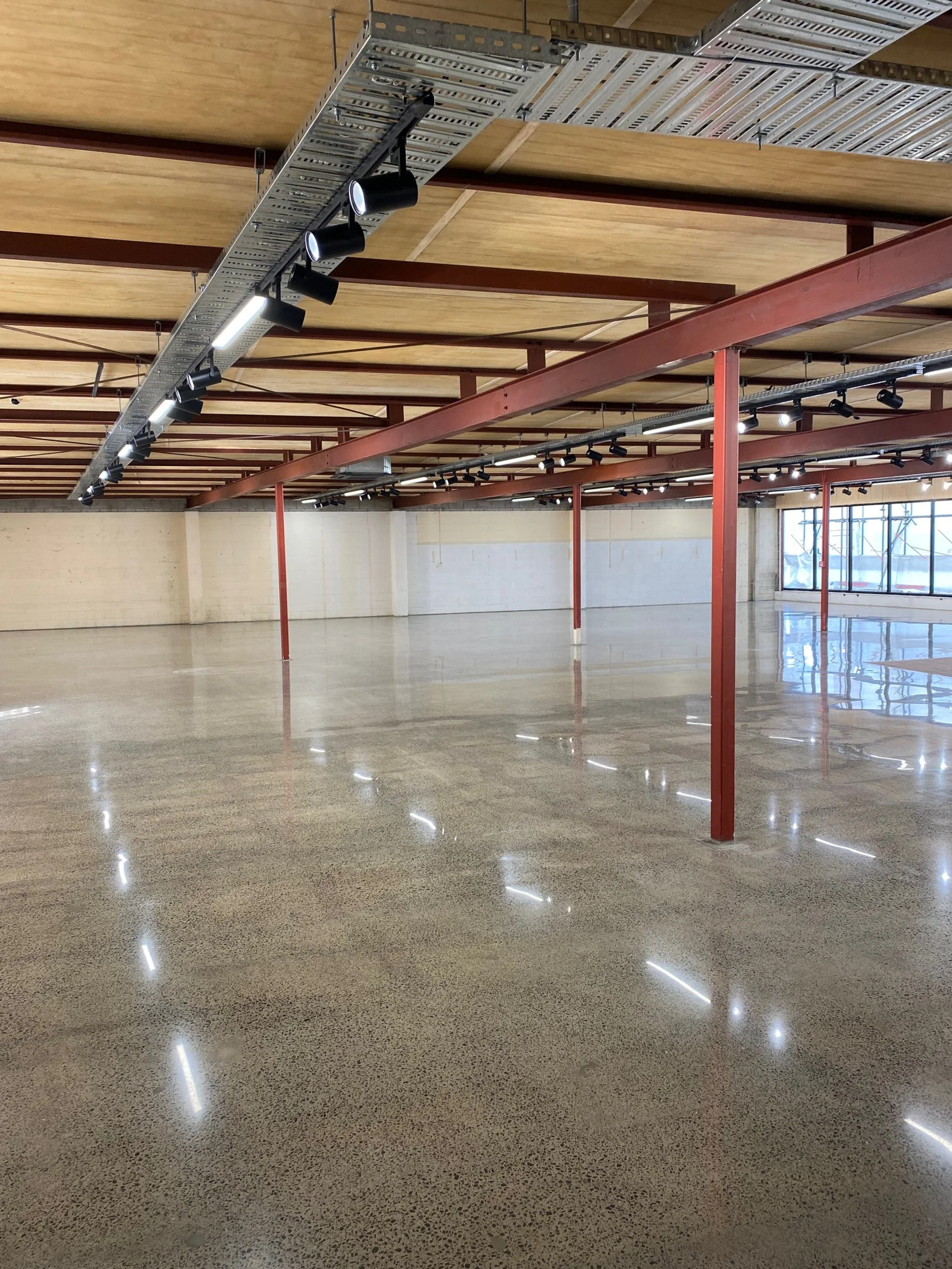 Empty indoor space with polished concrete floors, wooden ceiling with exposed beams, and large windows at the back.