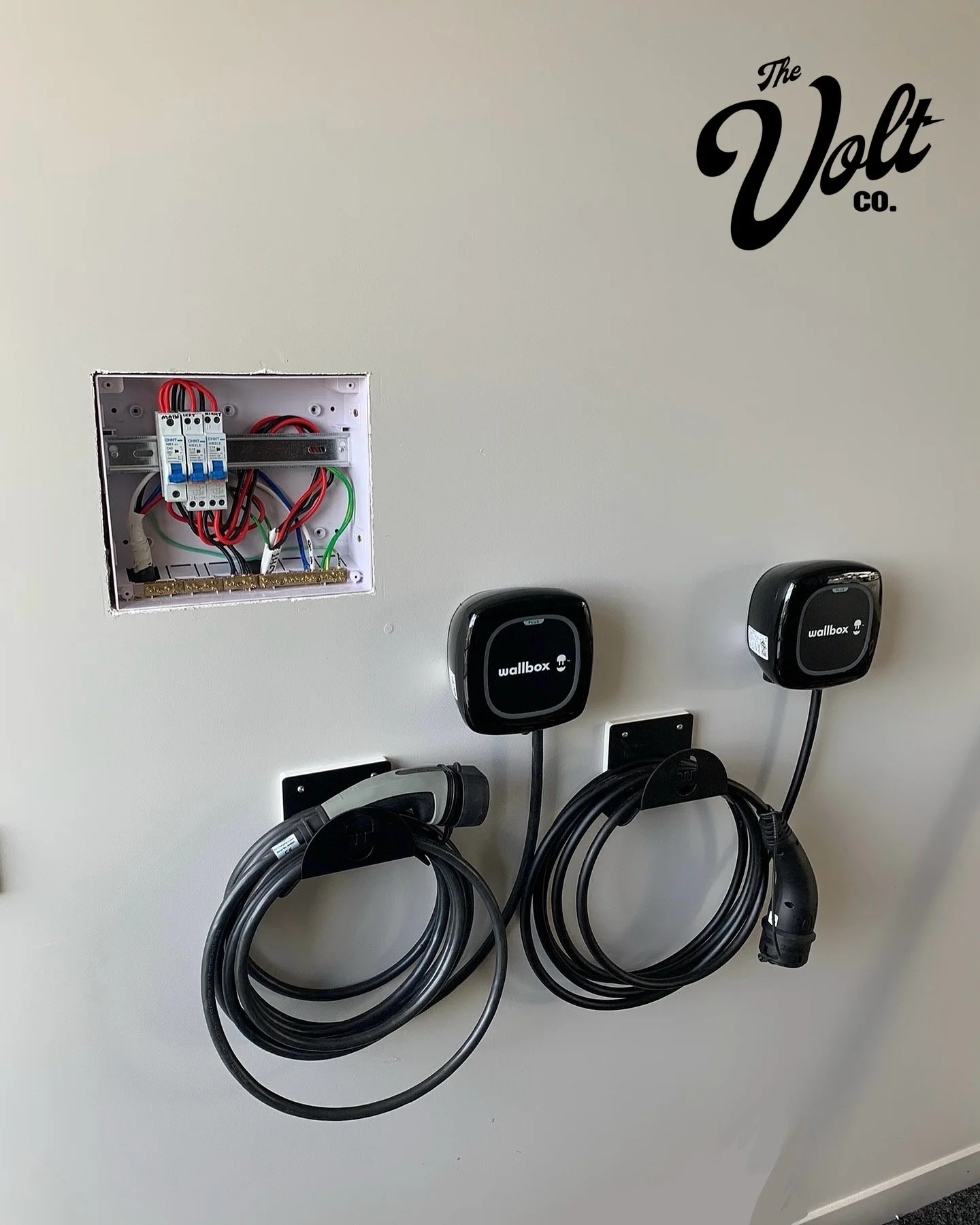 Electric vehicle charging station with two Wallbox chargers on a beige wall, and an open electrical box with wires.