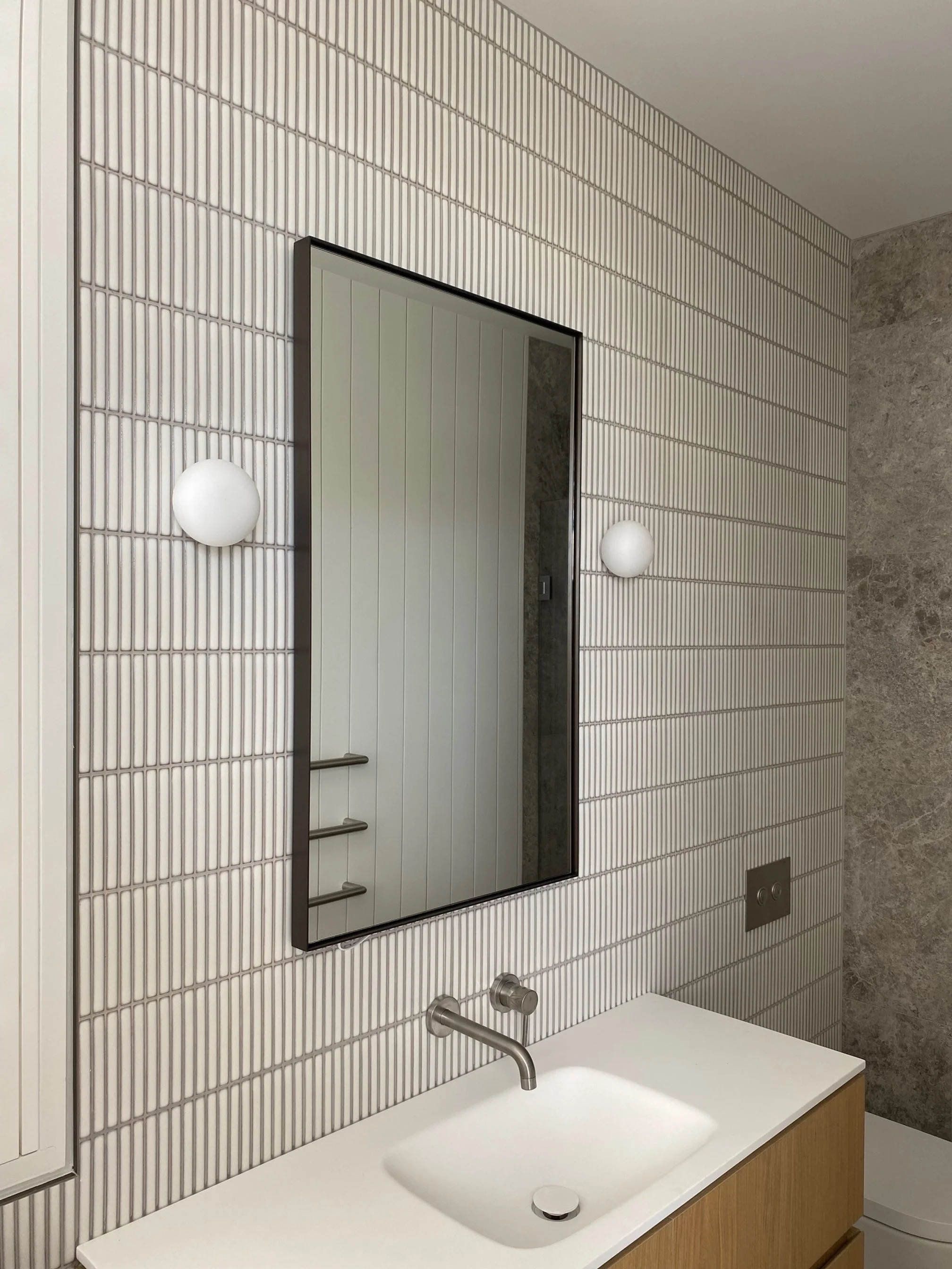 Modern bathroom with white tiled wall, a mirror, two globe wall lights, a wooden vanity with a white sink, and a textured gray wall.