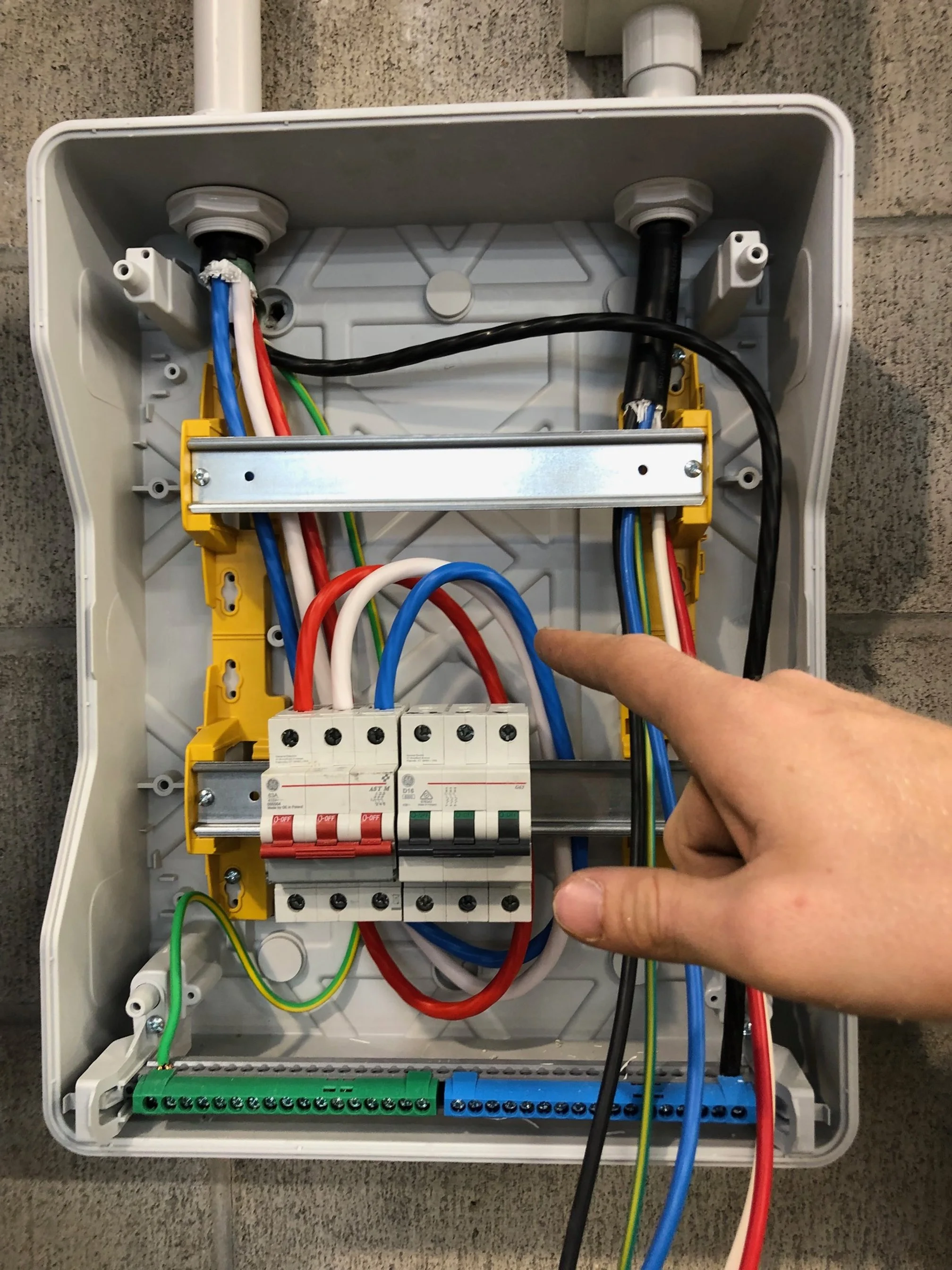 Open electrical breaker box with various wires and circuit breakers inside, and a person's finger pointing at one of the wires.