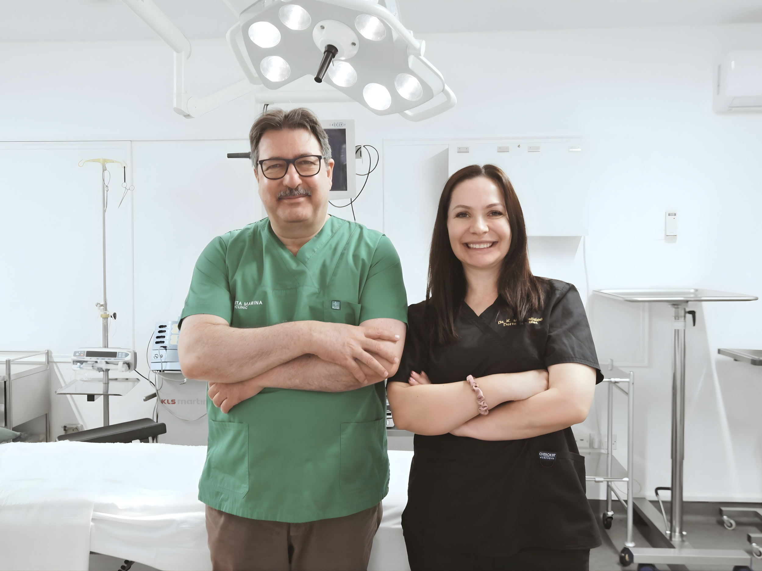 A male doctor in green scrubs and a female healthcare professional in black scrubs standing together in a hospital or medical room, smiling with arms crossed.
