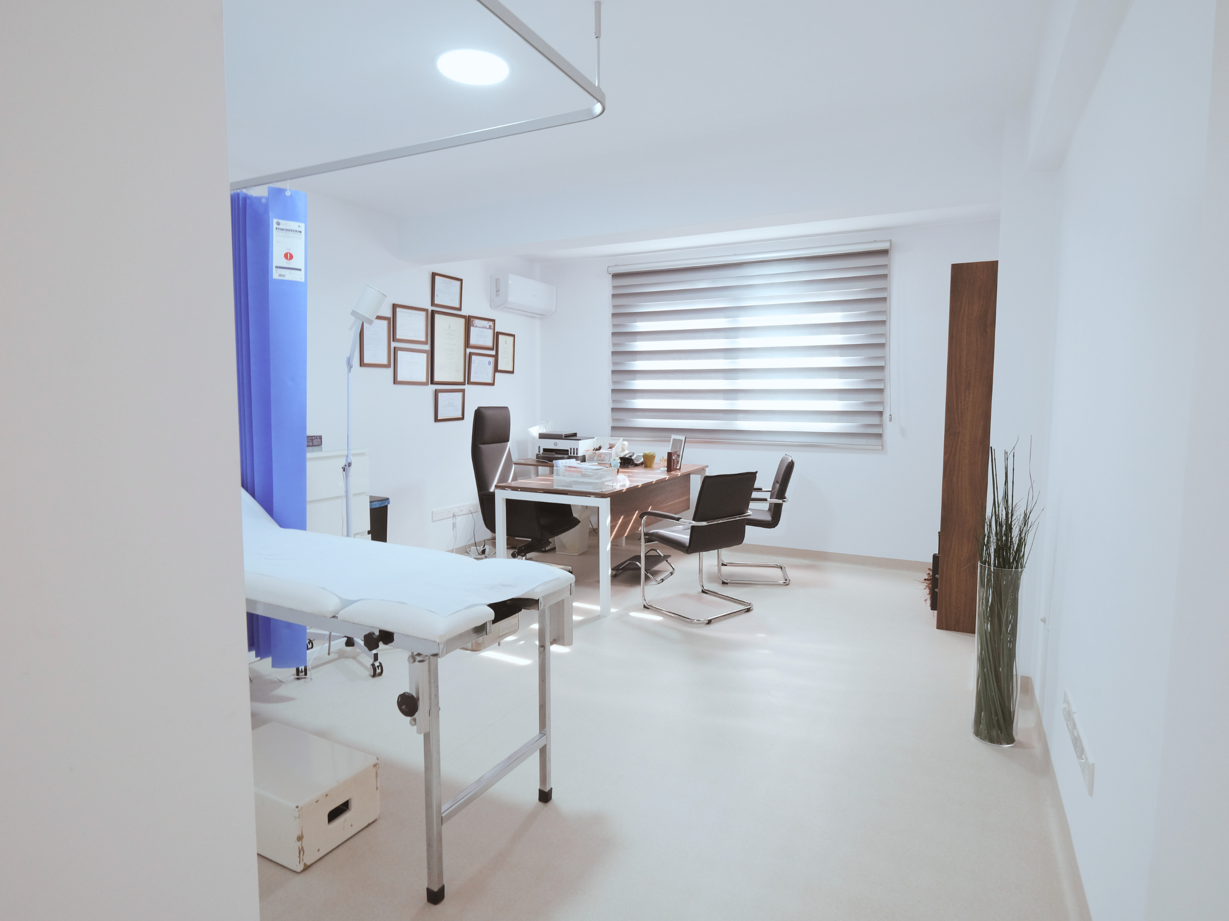 Santa Marina Polyclinic Paralimni Cyprus Empty medical examination room with patient bed, desk, chairs, certificates on wall, window with blinds, and potted plant.