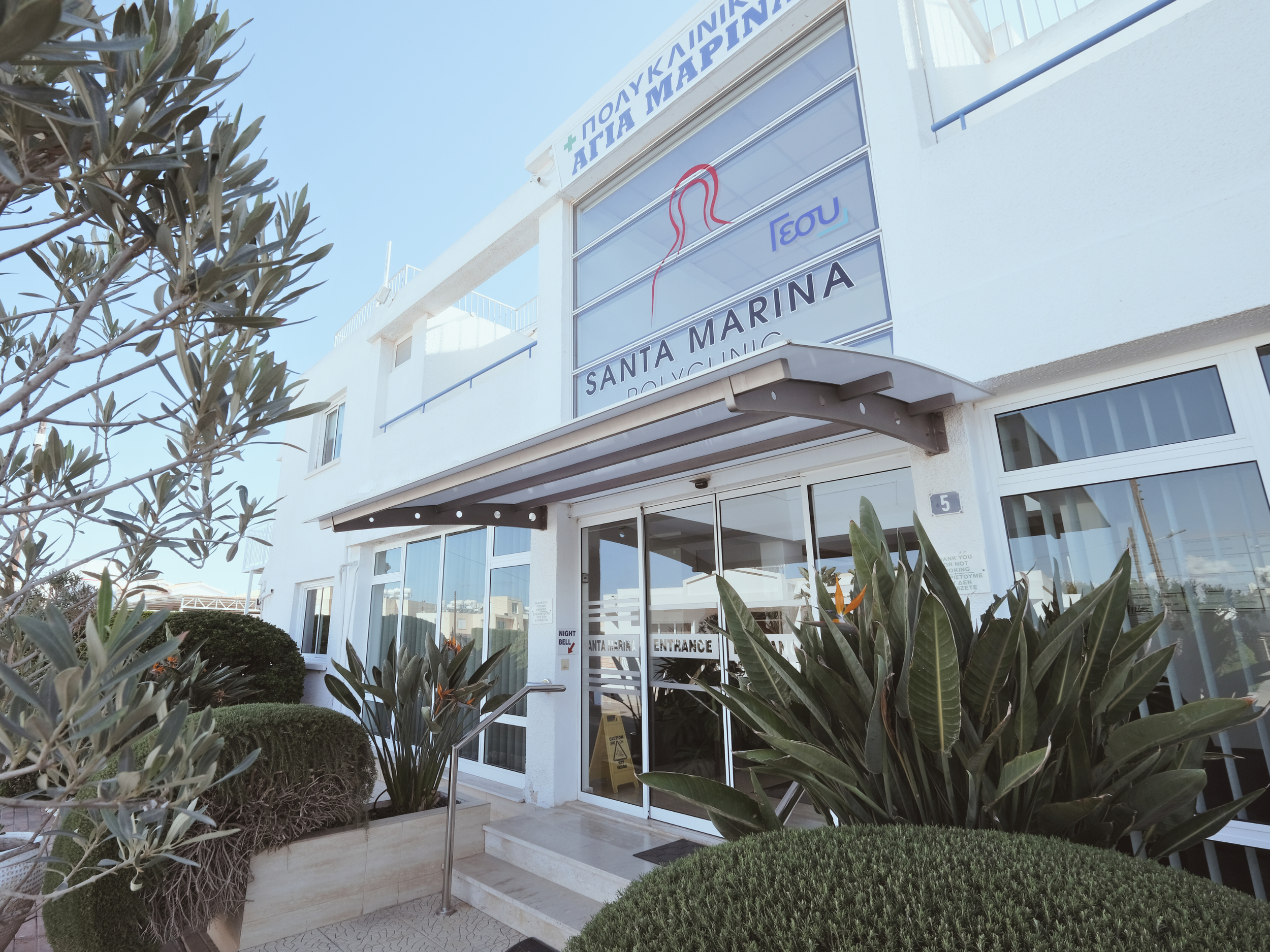 Exterior view of Santa Marina Polyclinic Paralimni Cyprus. a white building with glass doors, surrounded by green plants and bushes, with signs displaying the centre's name and logo.
