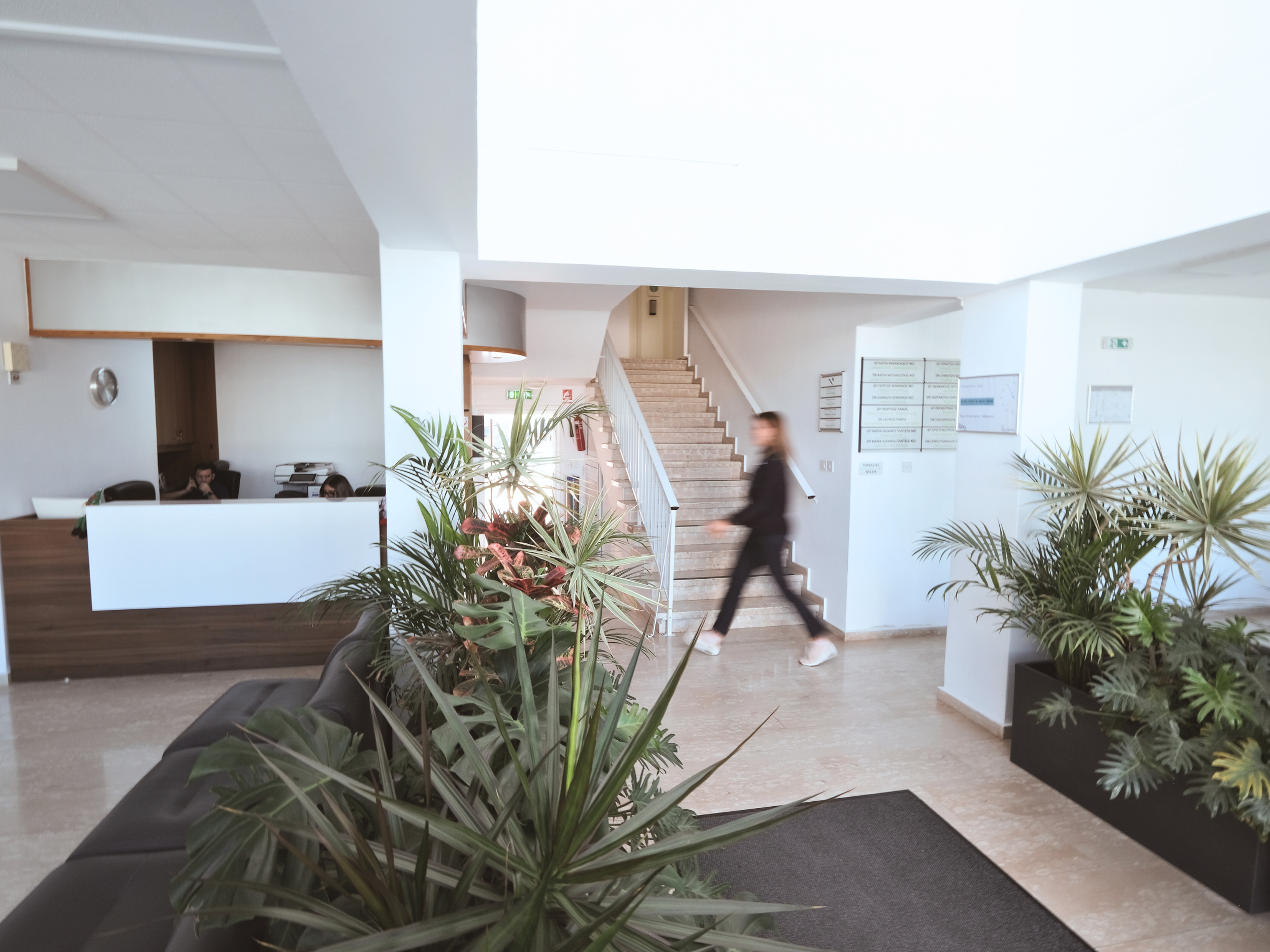 Santa Marina Polyclinic Paralimni Cyprus. Reception area hospital. A woman in black walking through a bright, modern office reception area with white walls, potted plants, a staircase, and a reception desk with staff.