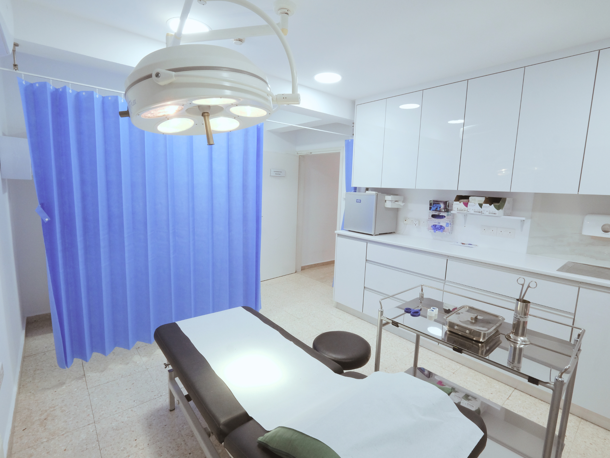 Santa Marina Polyclinic Paralimni Cyprus. Medical examination room with a bed, blue privacy curtain, overhead surgical light, and white cabinets.