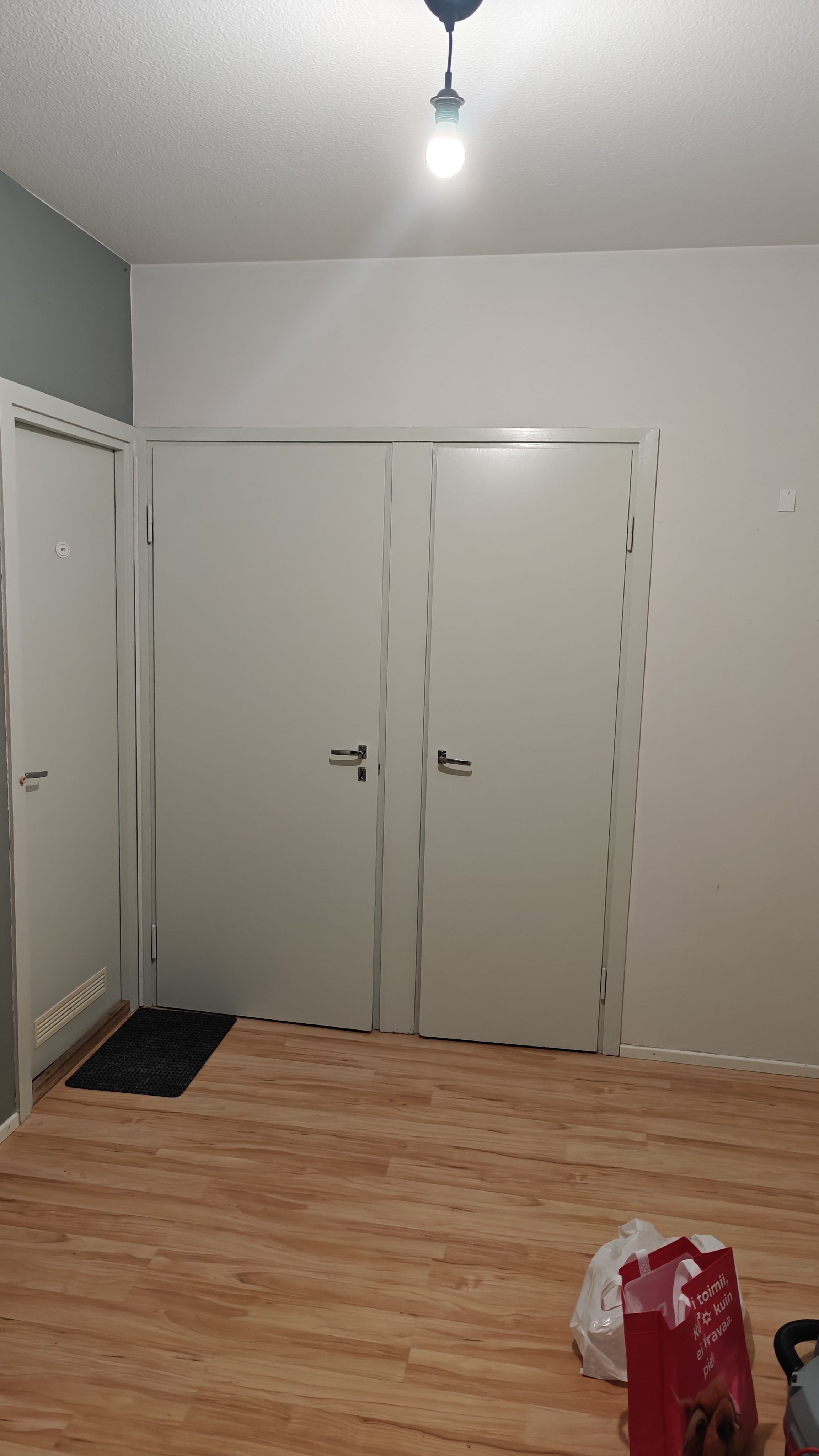 Kuvassa eteinen, jossa on kaksi valkoista kaappia, puiset lattiat ja valaisin katossa. 