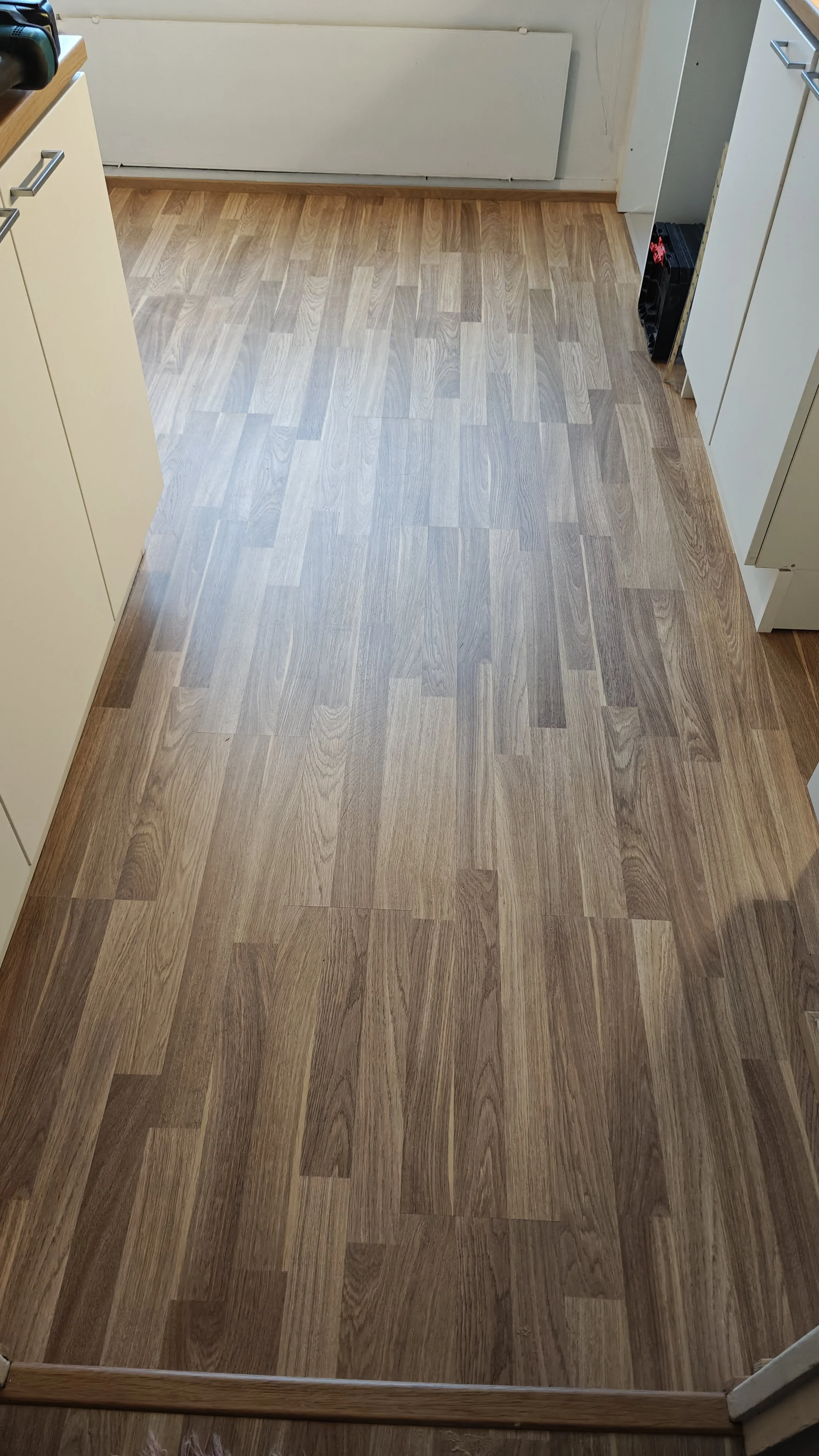 Kuvassa on puupintainen lattia ja osia keittiöstä, kuten kaappien ovia ja laatikoita, sekä osa keittiön seinää.