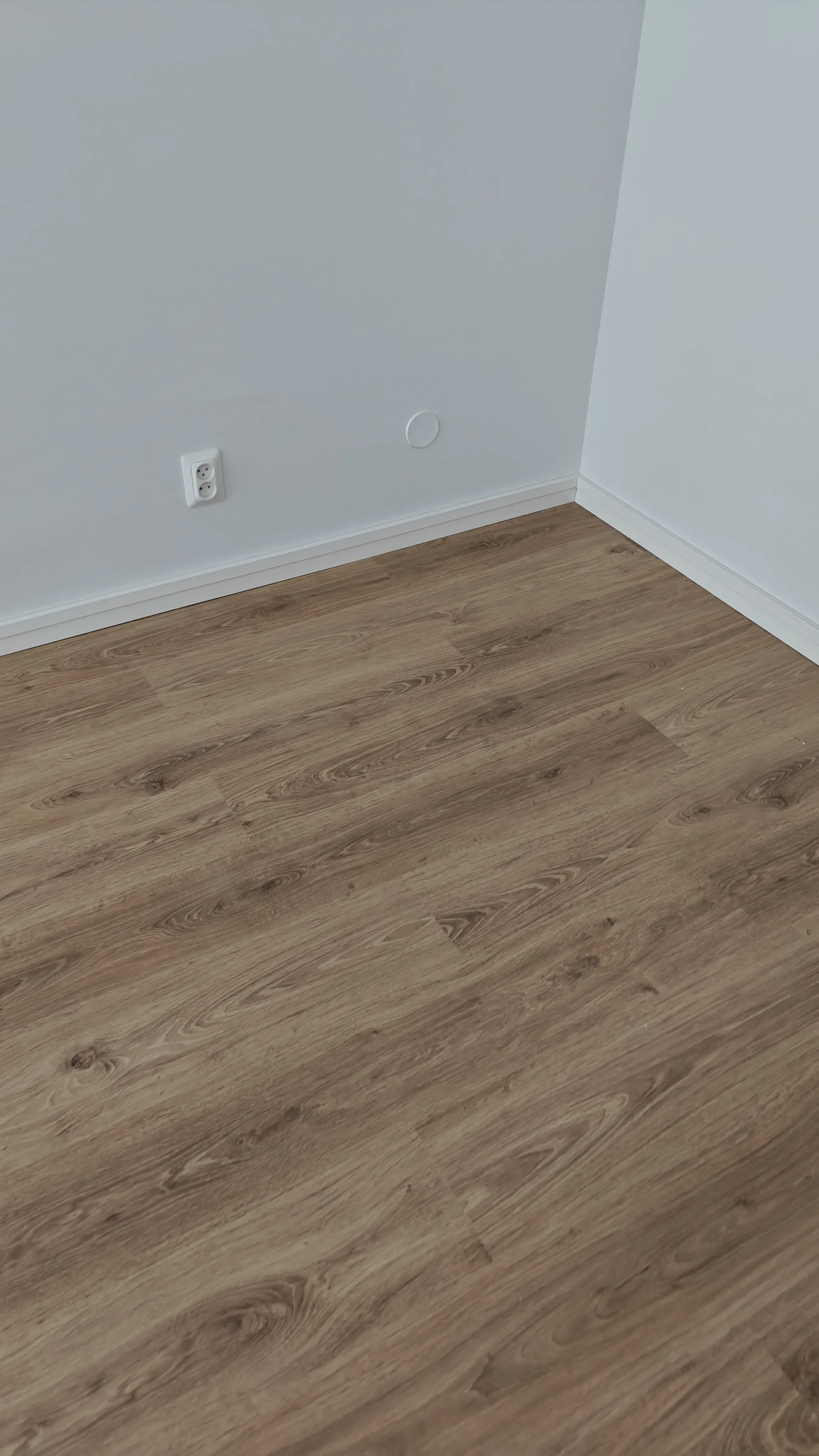 Kohde näyttää tyhjältä huoneelta, jossa on valkoinen seinä ja laminaattilattia. Seinällä on sähköpistoke ja kaksi pyöreää ilmanvaihtoventtiiliä.