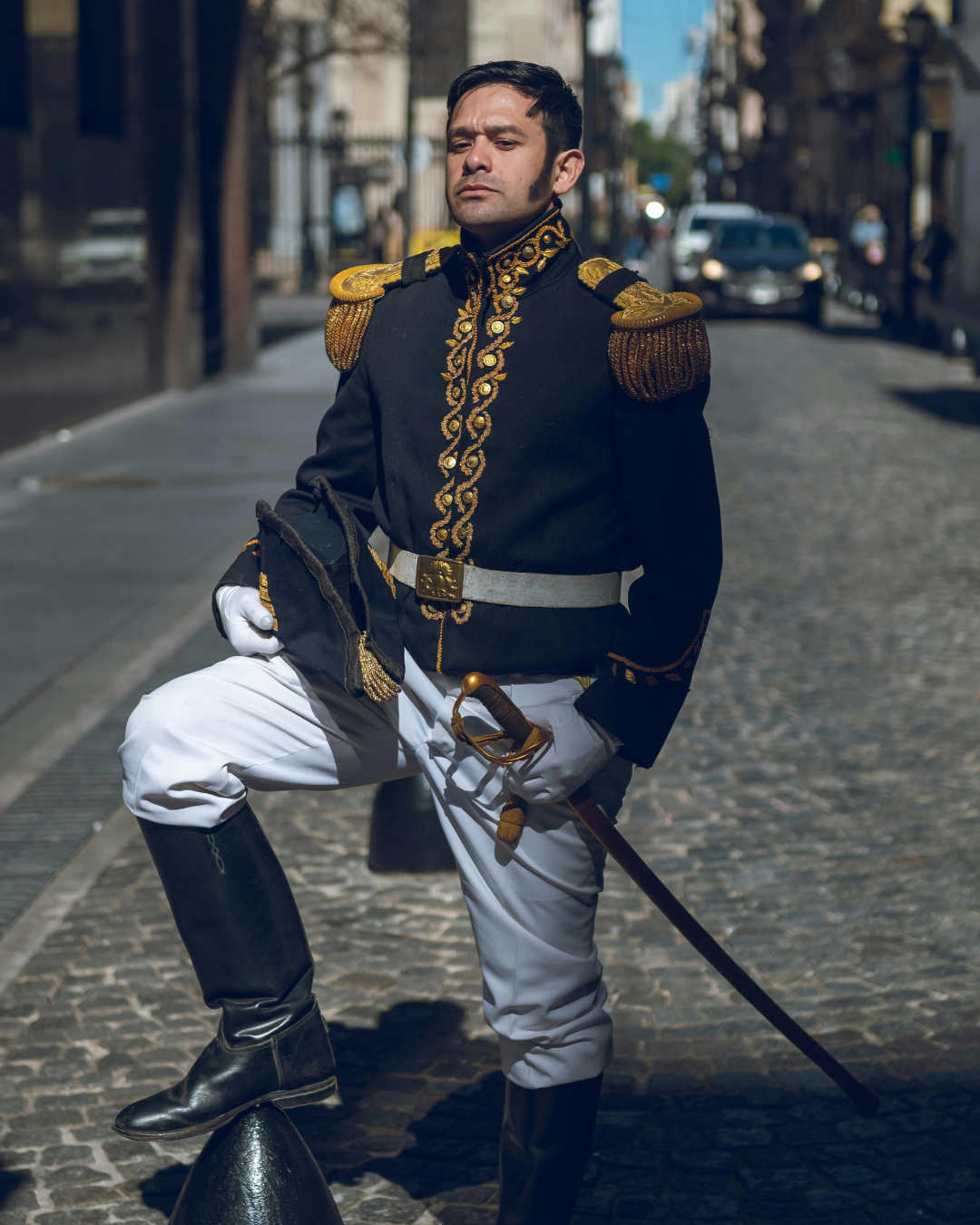 A man dressed in an ornate historical military uniform with gold embroidery, white pants, and black boots, holding a sword, standing on a cobblestone street with modern cars and urban buildings in the background.