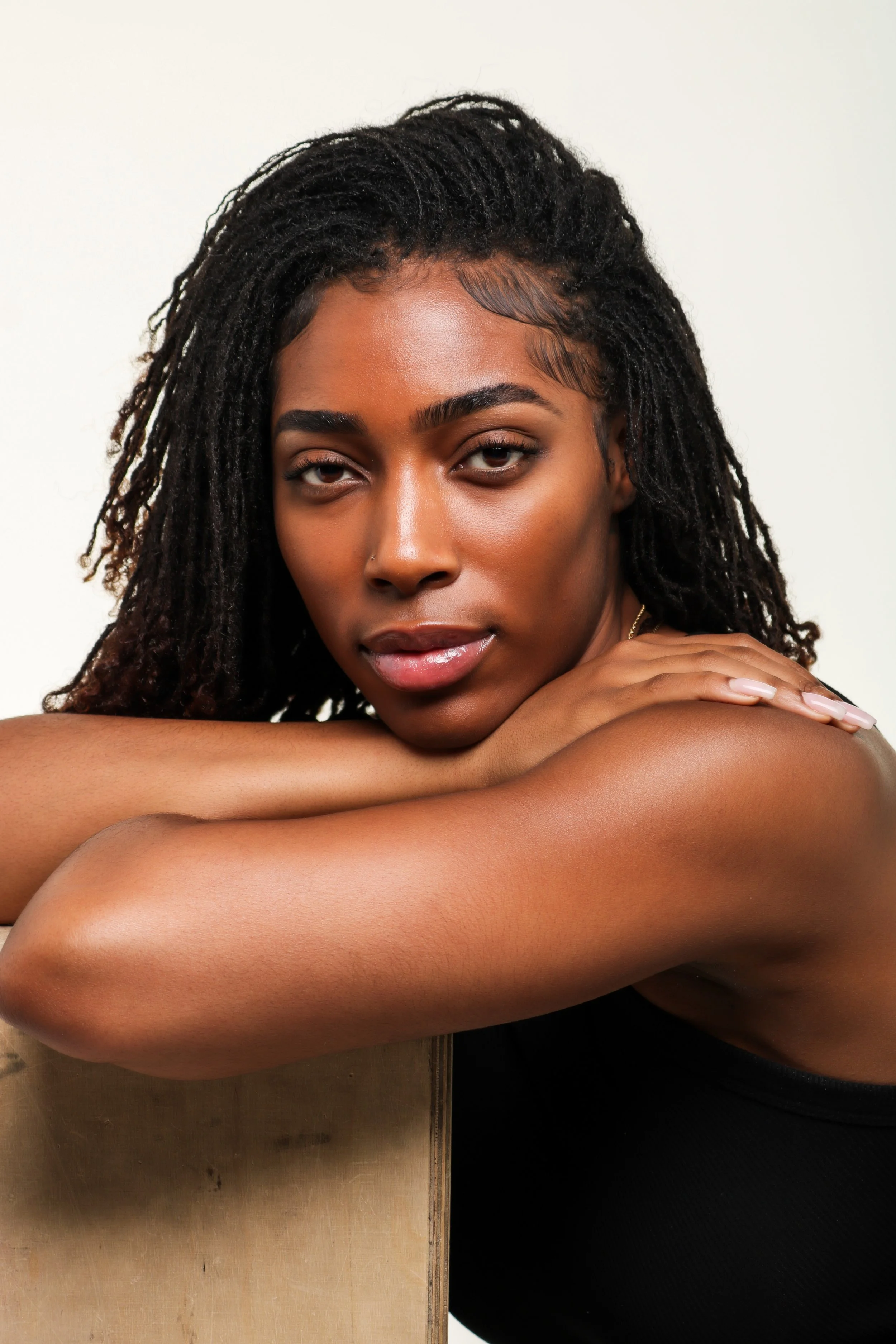 A young woman with dark, curly hair and a nose piercing rests her arms on a wooden surface, looking at the camera with a relaxed expression.