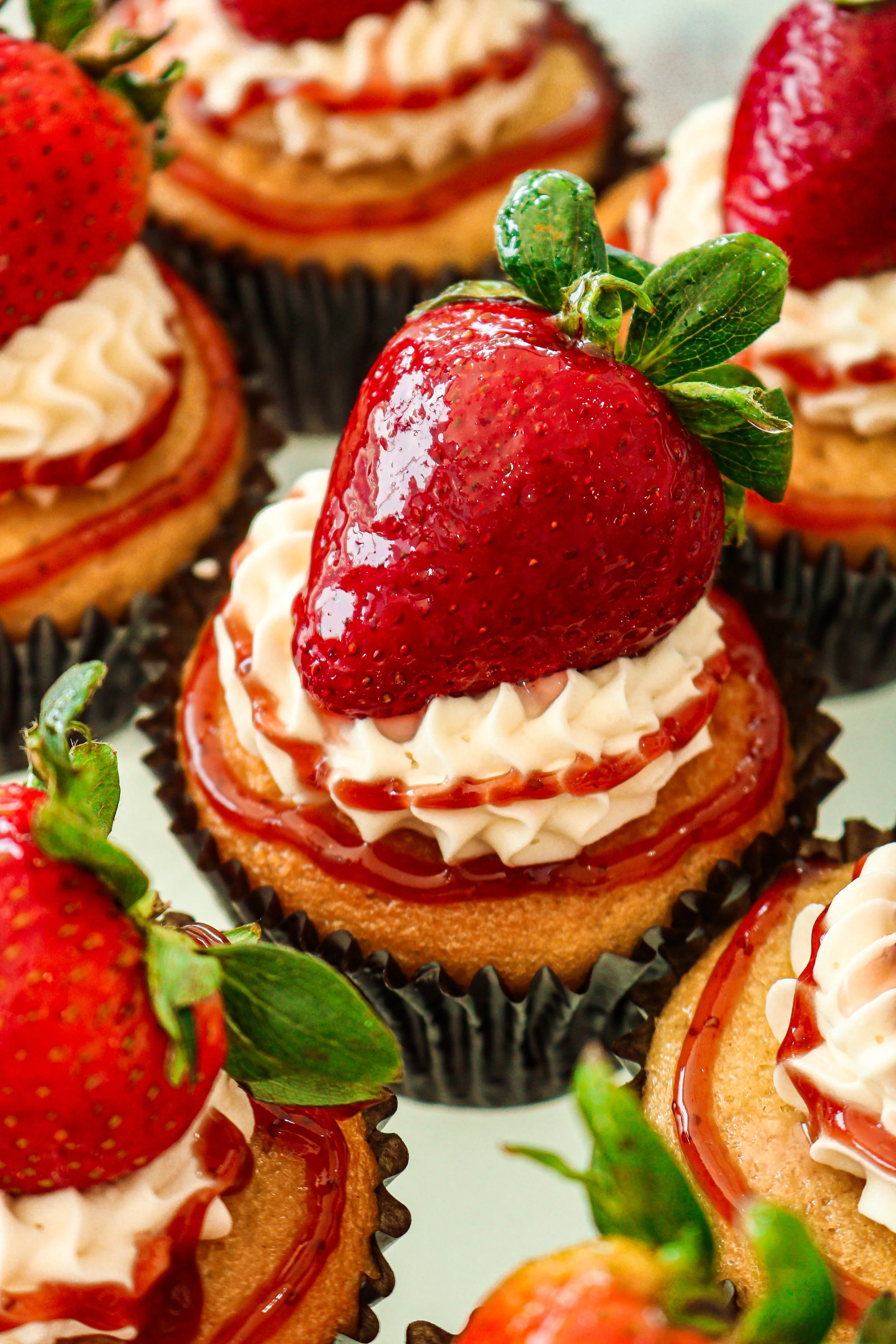 Close-up of strawberry-topped cupcakes with frosting and caramel drizzle, in black cupcake liners.