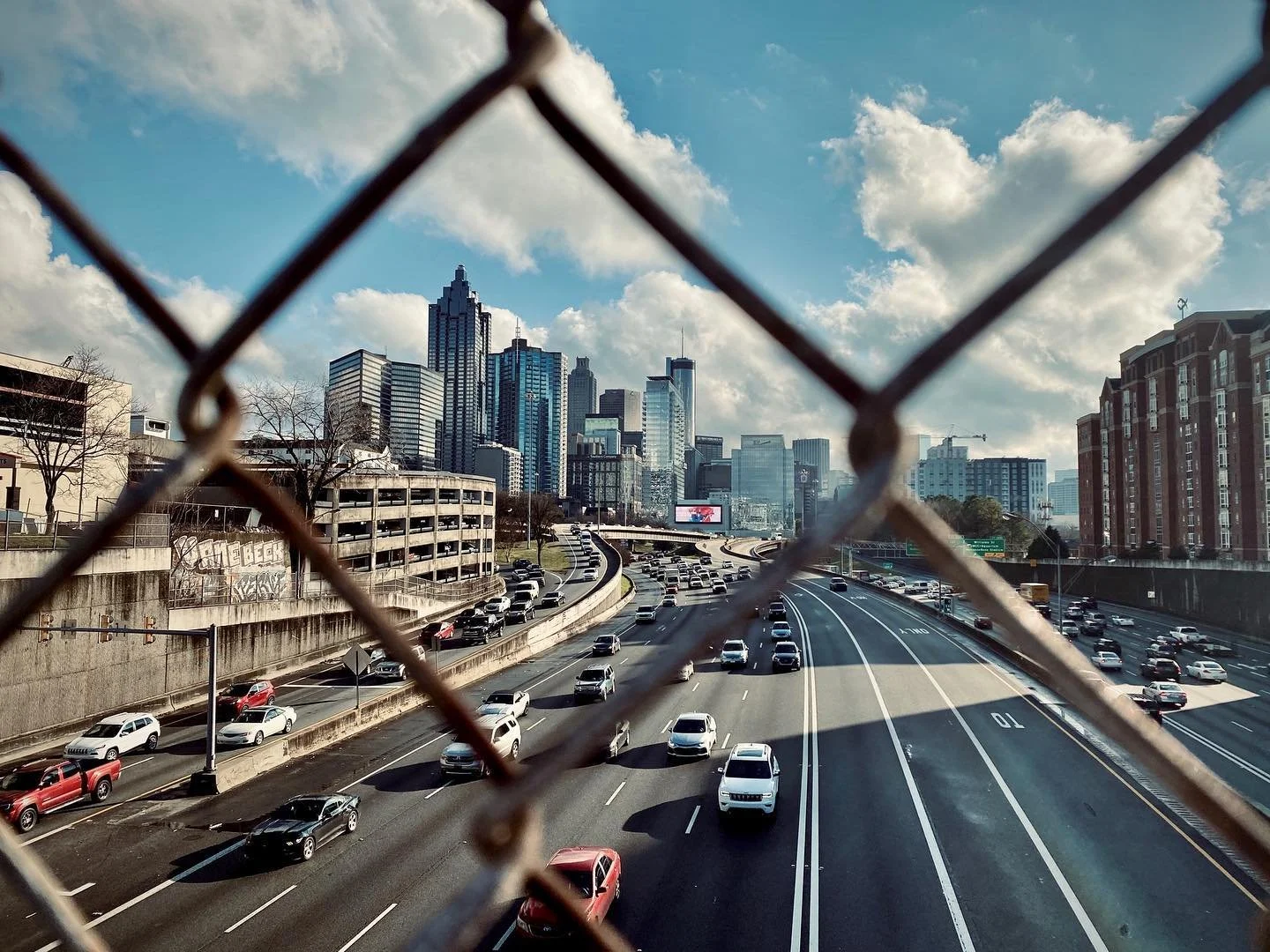 City skyline with skyscrapers seen through a chain-link fence over a busy highway with moving cars and trucks under a partly cloudy sky.