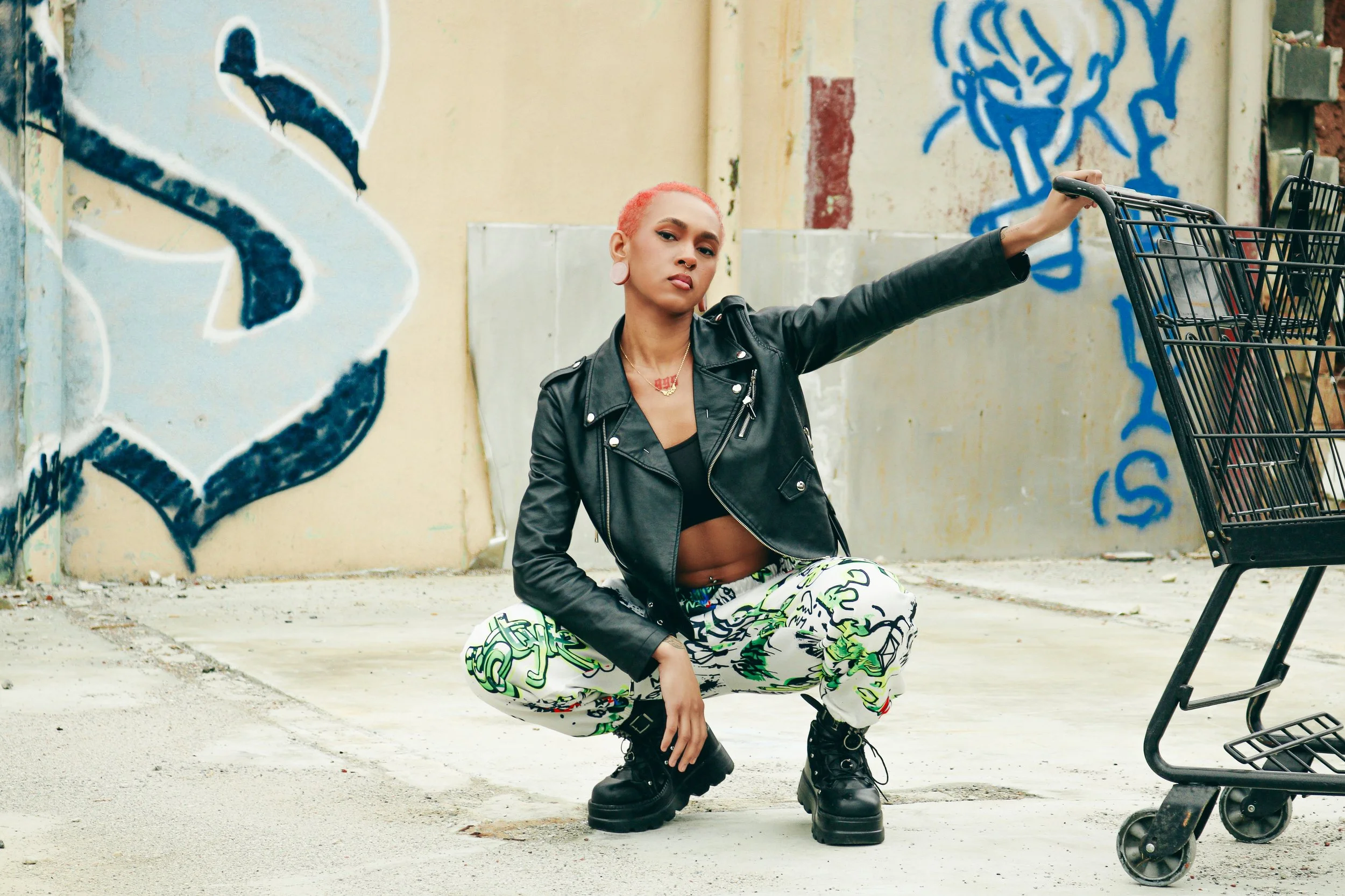 A woman with short pink hair squats on a city sidewalk, wearing a black leather jacket, black top, patterned pants, and black boots, next to an empty shopping cart, with a graffiti-covered wall behind her.