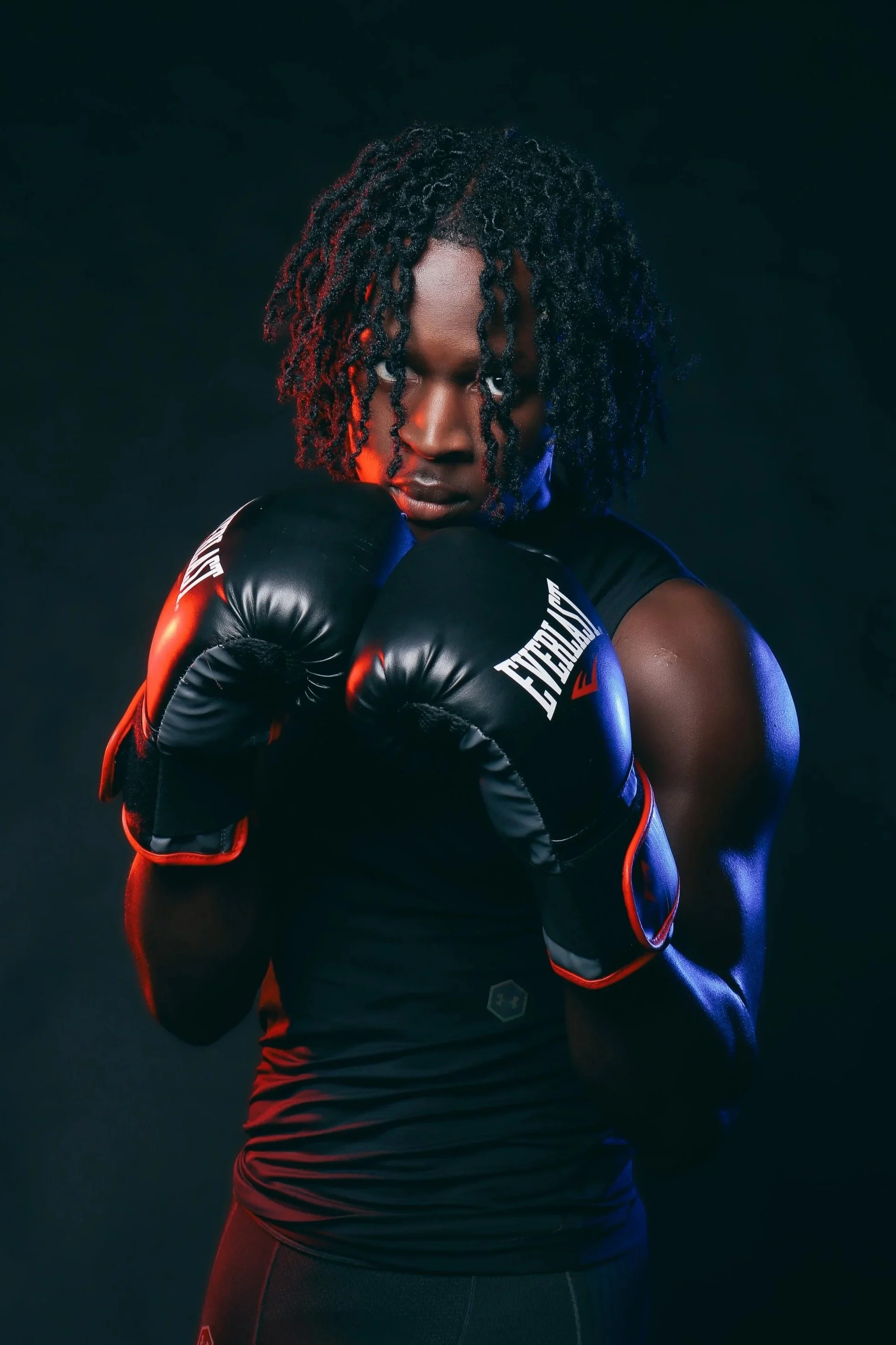 A man with curly hair wearing black boxing gloves and athletic wear, posing in a boxing stance against a dark background.