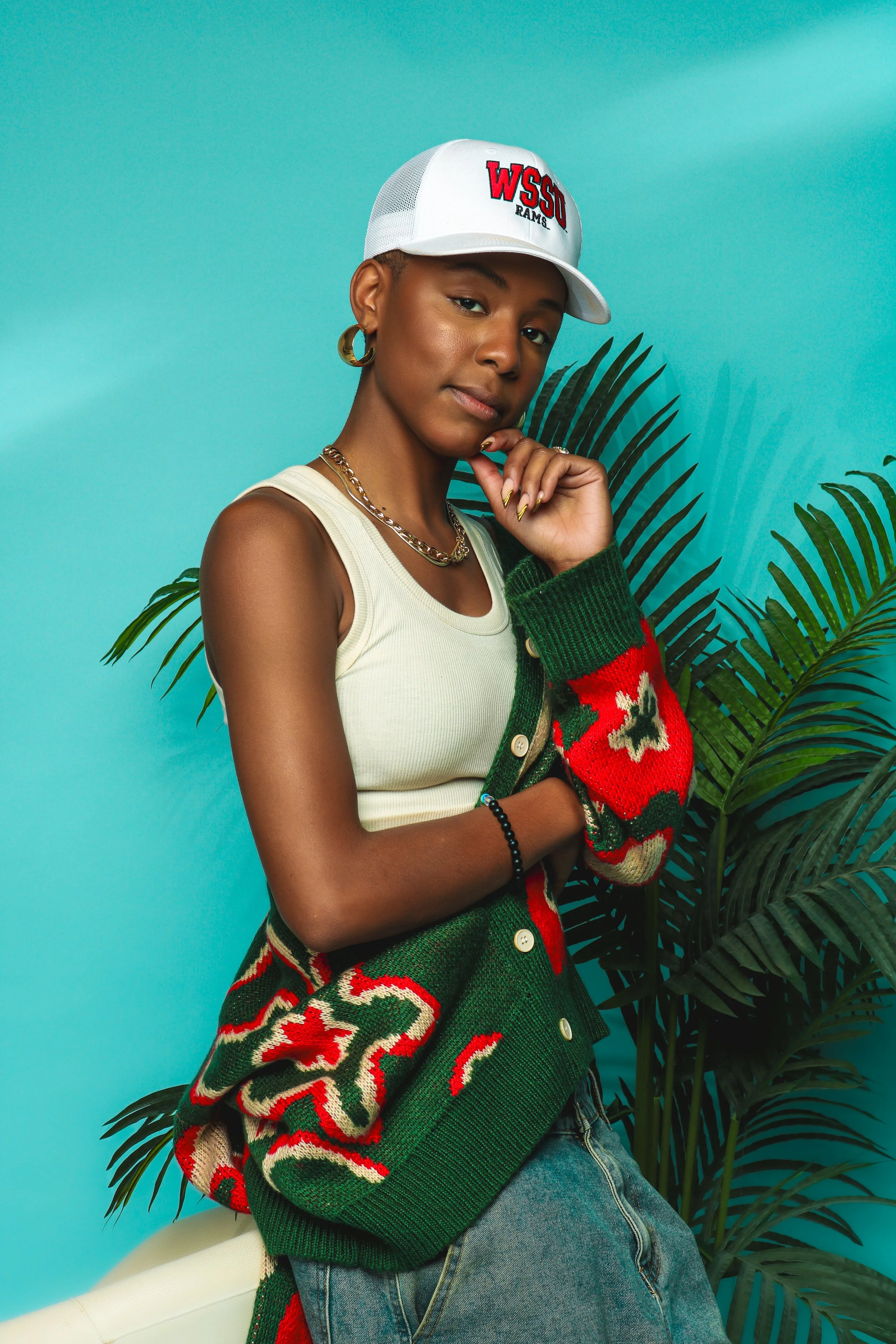A young woman with short hair, wearing a white cap with red and black embroidered text 'WSU RAMS', gold hoop earrings, gold chain necklace, a cream spaghetti strap top, a green cardigan with red and white floral pattern draped over her arms, and blue jeans. She is standing beside a large green plant with a vibrant turquoise background.