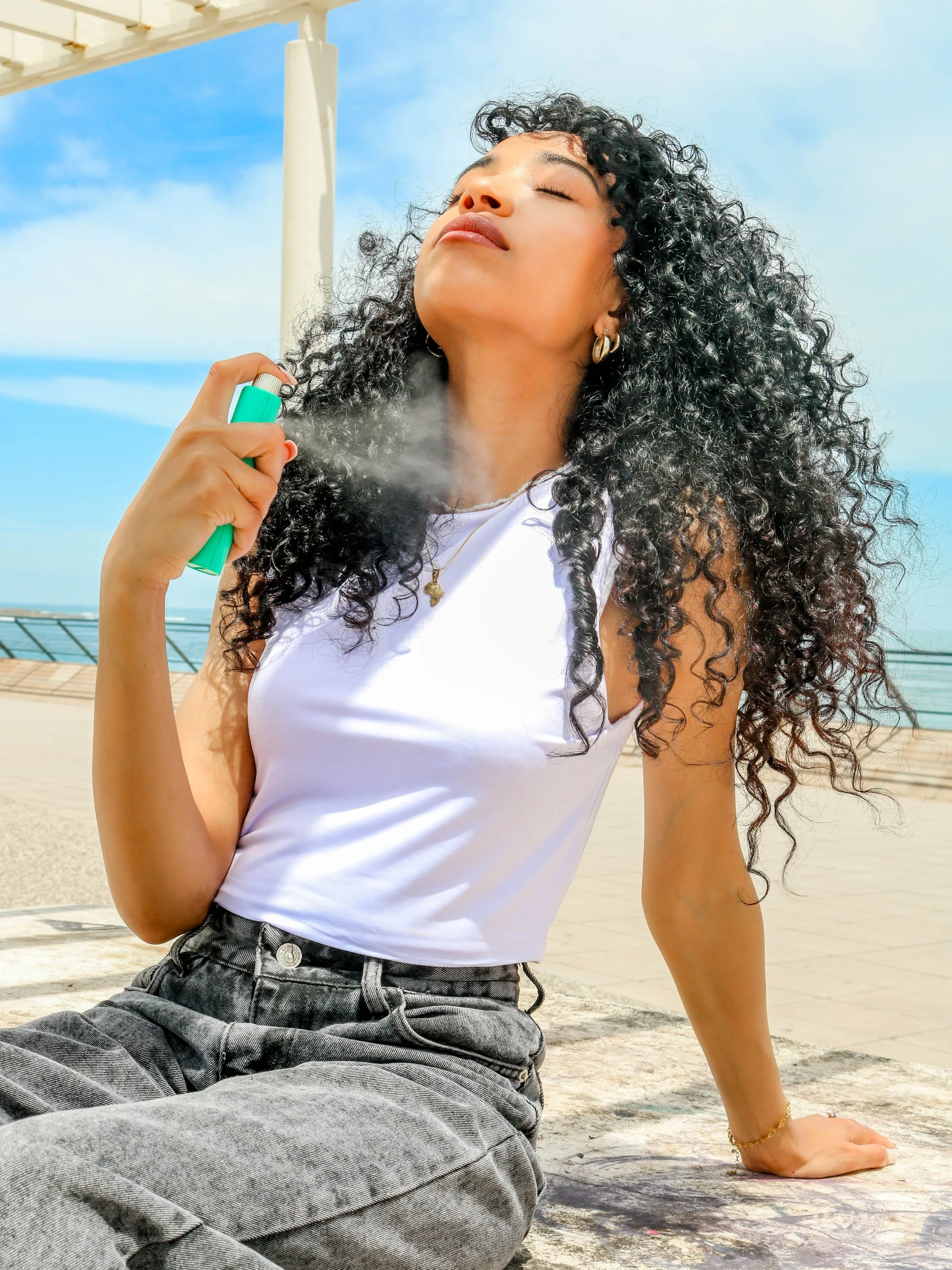 A young woman with curly black hair is spraying perfume on her skin outdoors near the beach, with her eyes closed and a relaxed expression.