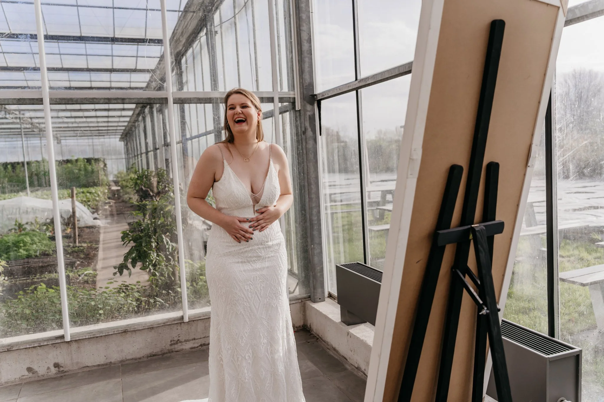 Vrouw in witte trouwjurk lacht in een kas met planten en grote ramen.