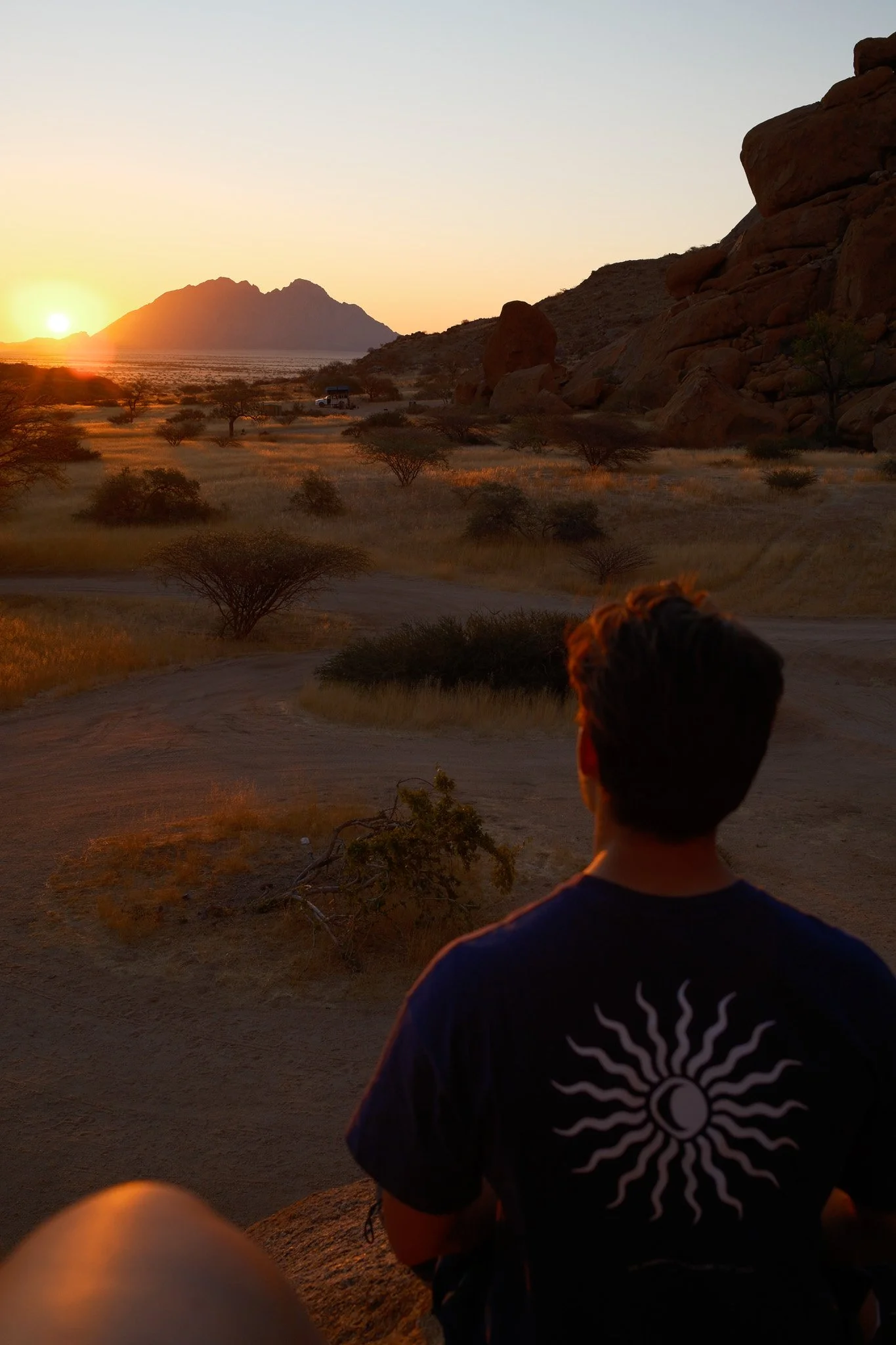 Een jongen zit op een rots en kijkt naar een zonsondergang over een woestijnachtig landschap met rotsen, kleine struiken en een wandeling in de verte.