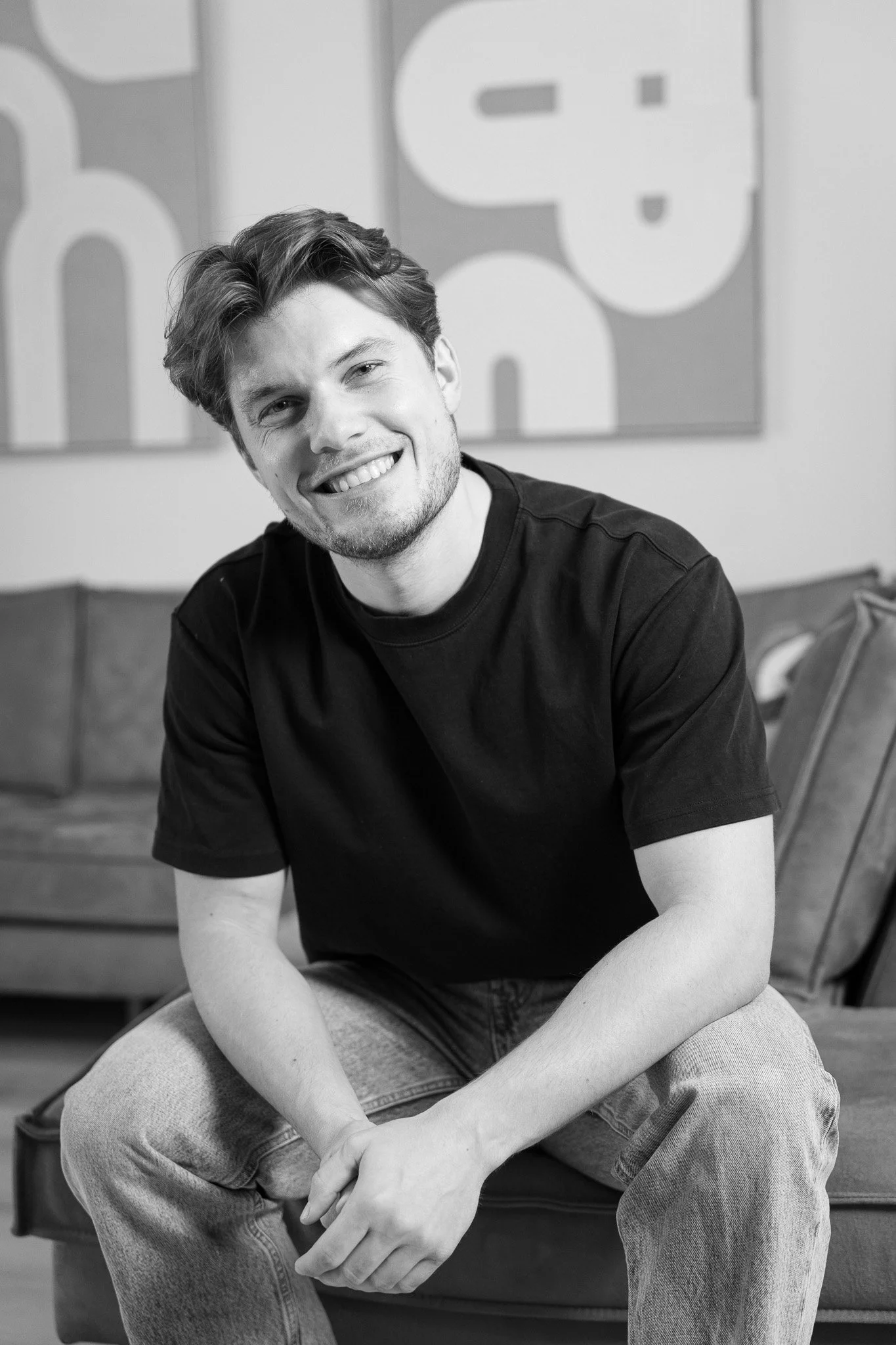 Een lachende jonge man met kort haar, zittend op een bank in een interieur met moderne kunst aan de muur, in zwart-wit.