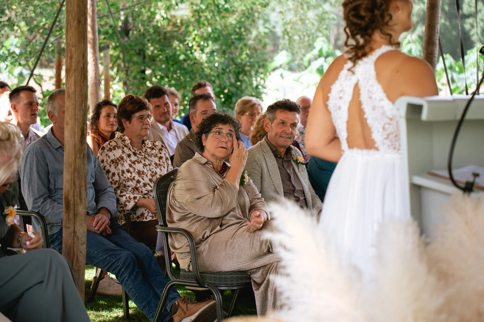 Een groep mensen zit in een groene, openlucht omgeving en kijkt naar een vrouw die voor hen spreekt. De vrouw draagt een witte jurk met lace details op de rug, ter gelegenheid van een huwelijk.