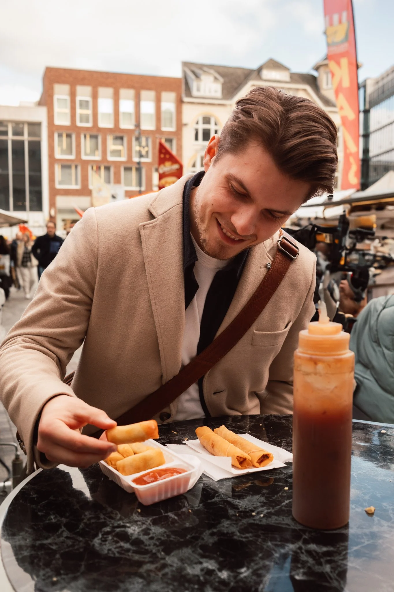 Een lachende man eet een lenterol met saus bij een tafel buiten, met gebouwen en menigten op de achtergrond.