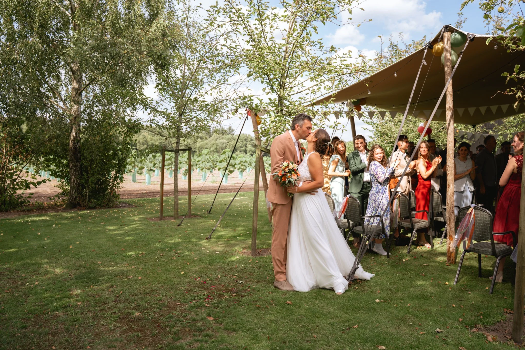 Trouwkoppel kust elkaar na de ceremonie onder een tent buiten, omringd door gasten die applaudisseren en lachen, met een achtergrond van bomen en een blauwe lucht.