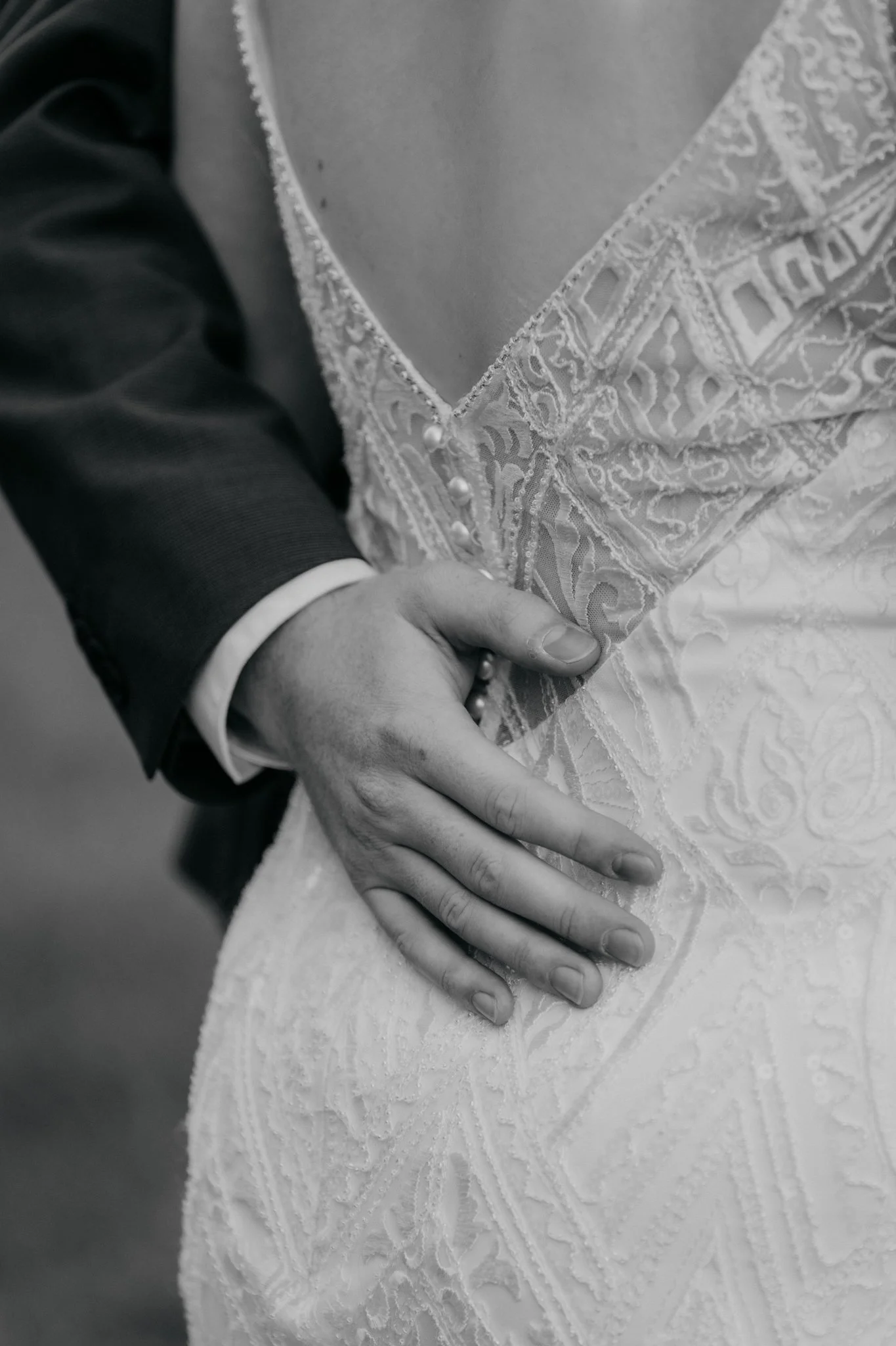 Close-up van een paar handen, een wijd open en de andere hand rust op de heup, tijdens een bruiloft. In zwart-wit, met een trouwjurk en een pak zichtbaar.