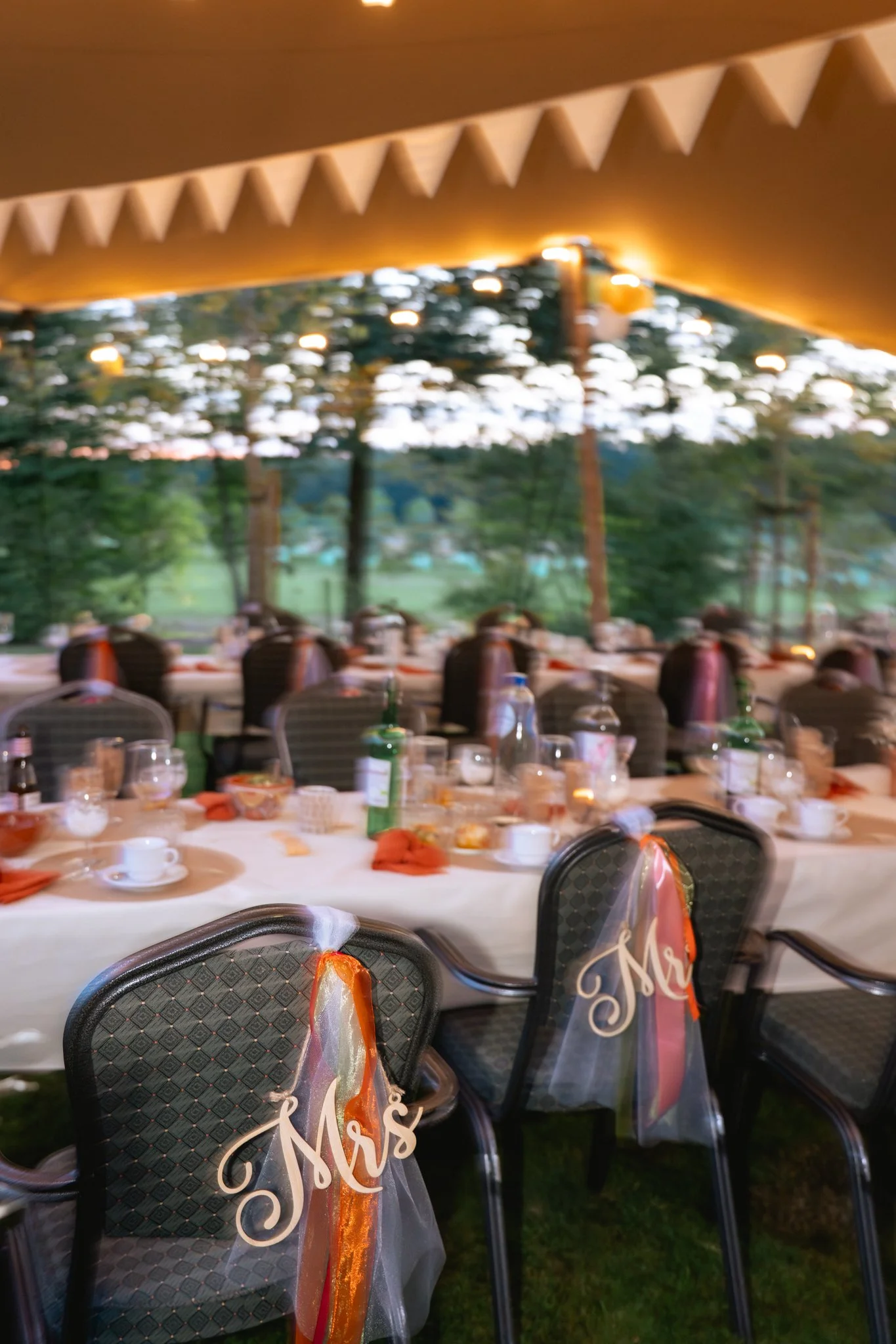 Decoratie voor een bruiloft met stoelen die 'Mrs' en 'Mr' labels hebben, een feesttent met verlichting en een tafel gedekt voor een trouwfeest, buitenomgeving met bomen.