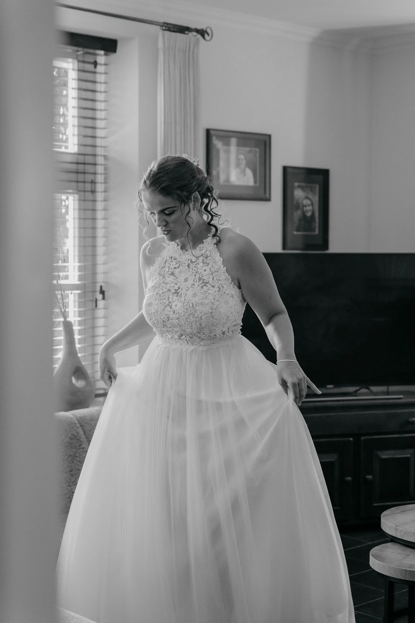 Een bruid in een trouwjurk die haar jurk vasthoudt, binnen in een kamer met foto’s aan de muur.