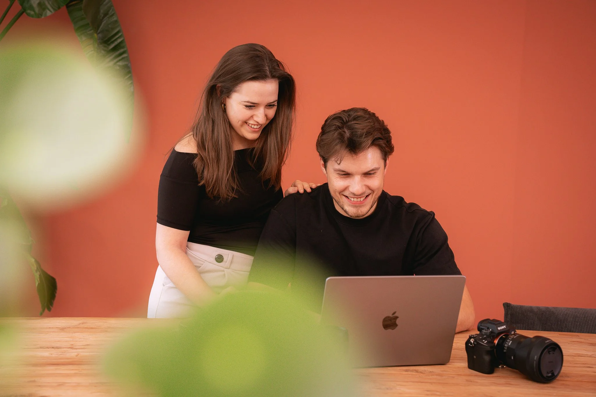 Twee fotografen werken samen op een laptop op een houten tafel, met een camera ernaast. Ze glimlachen en hebben plezier.