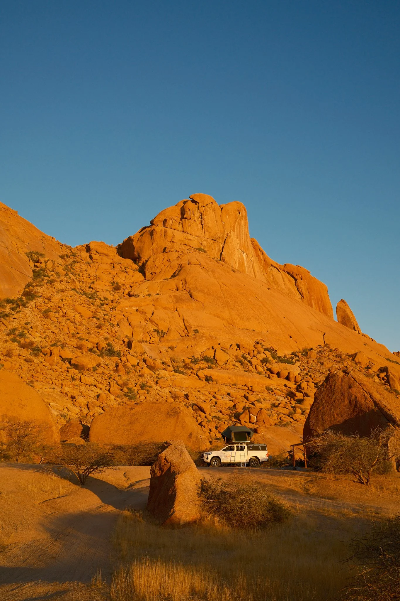 Een witte jeep met een daktent rijdt door een desert landschap met grote rotsen onder een blauwe hemel.