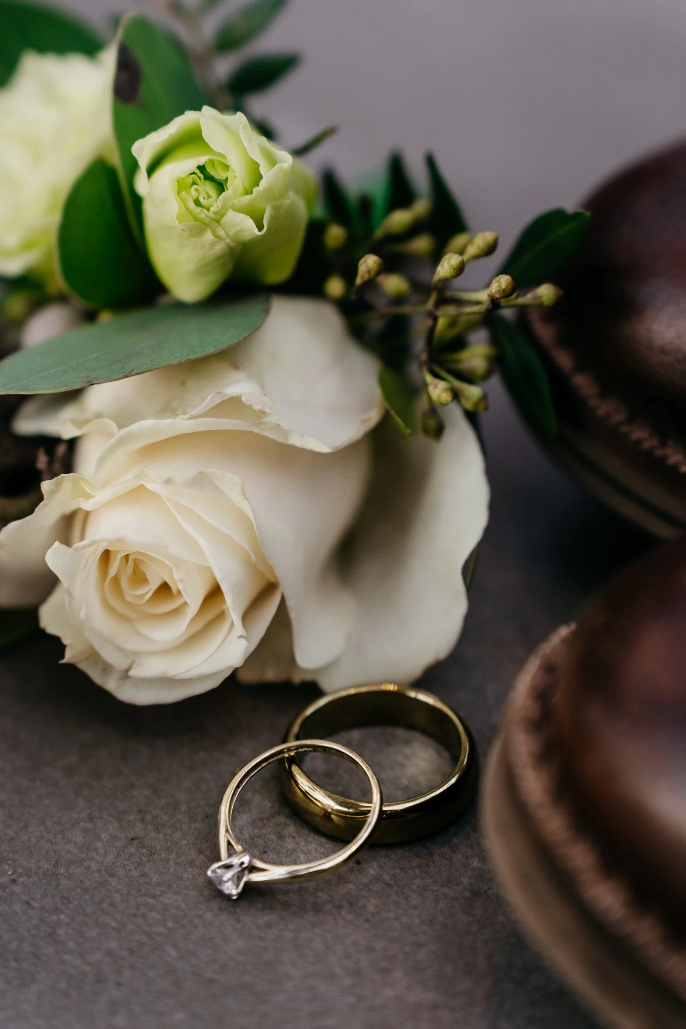 Detailfoto van een huwelijk-ring met diamant ligt naast een gouden trouwring, met witte rozen, groen blad en schoenen op een donkere achtergrond.